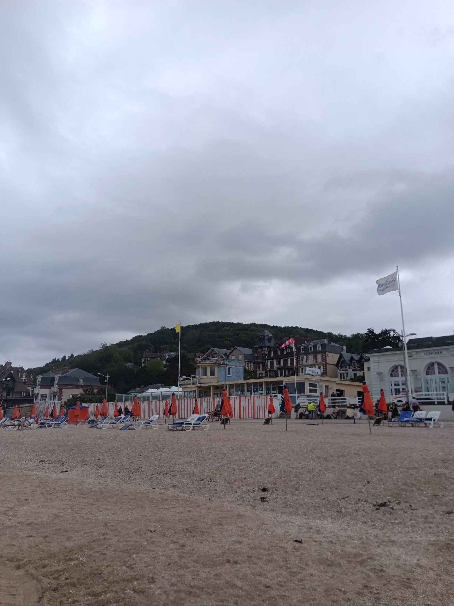 The image depicts a beach in Houlgate, France, on a cloudy day. In the foreground, a pebbly beach stretches across the frame, with a few dark patches suggesting seaweed or debris. Rows of empty deck chairs and closed orange umbrellas are arranged on the sand, indicating a lack of beachgoers or the early/late hours of operation.
In the middle ground, a line of buildings forms the backdrop to the beach. These buildings appear to be a mix of architectural styles, with some featuring half-timbered elements and others presenting more modern facades. A building on the right side of the frame is labeled "CINEMA." Other structures include a white building with arched windows and a long, glass-fronted enclosure with orange vertical stripes.
Further back, a lush green hillside is covered with trees and dotted with houses. A single yellow flag flies from a flagpole on the hill. The sky is overcast with heavy grey clouds, casting a subdued light over the scene. A flag with a logo, possibly a business or local emblem, is visible on a flagpole in front of the cinema building. A few individuals are scattered throughout the middle ground, near the buildings and seating areas, some sitting on chairs or standing in small groups. One person is seen walking a dog on the beach.