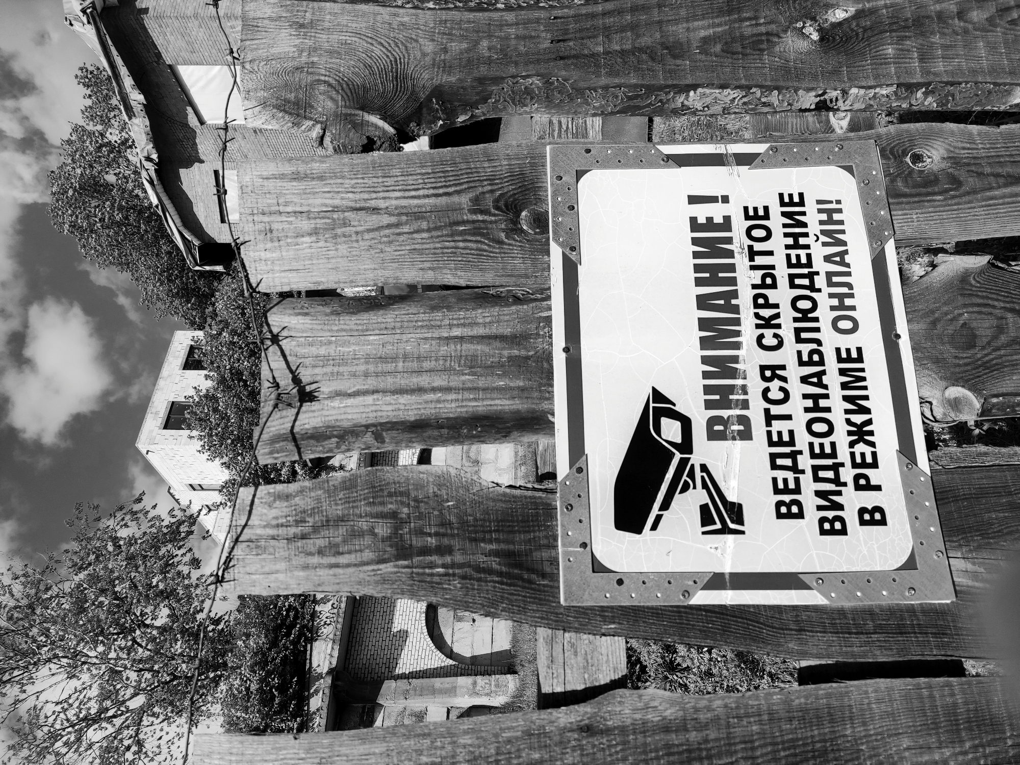 Captured in black and white, the image presents a view of a wooden fence, angled towards the sky. Attached to the fence is a sign in Russian, featuring a security camera icon and the text "ВНИМАНИЕ! ВЕДЕТСЯ СКРЫТОЕ ВИДЕОНАБЛЮДЕНИЕ В РЕЖИМЕ ОНЛАЙН!", which translates to "WARNING! HIDDEN VIDEO SURVEILLANCE IS IN PROGRESS ONLINE!". Barbed wire runs along the top of the fence. Behind the fence, partially obscured by foliage, a building with brickwork and windows is visible. Based on the architecture and the language on the sign, this image likely originates from the city of Hrodna, Belarus.