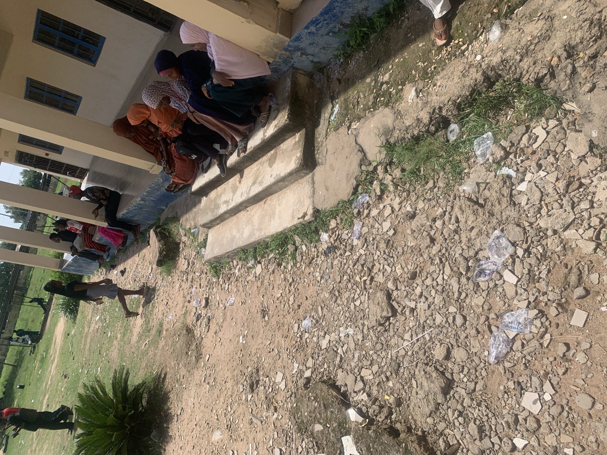 The image depicts a group of people, predominantly women wearing hijabs, sitting on steps outside a building. Some are looking down, possibly engaged in conversation or a shared activity. The building appears to be a school or a community center, with blue-framed windows visible on the upper floor. The ground is covered with gravel, dirt, and scattered pieces of debris, suggesting an unpaved outdoor area.
In the foreground, a person is seen walking away from the camera, dressed in casual attire. The overall scene suggests a daytime setting, likely during a period of rest or a break. The atmosphere seems relaxed, with individuals casually gathered on the steps.
Notable details include the presence of plastic water bottles strewn on the ground, indicating littering or a recent event where water was distributed. The location appears to be Jalingo, Nigeria, a context that might imply a warm, sunny climate. There is no visible text in the image.