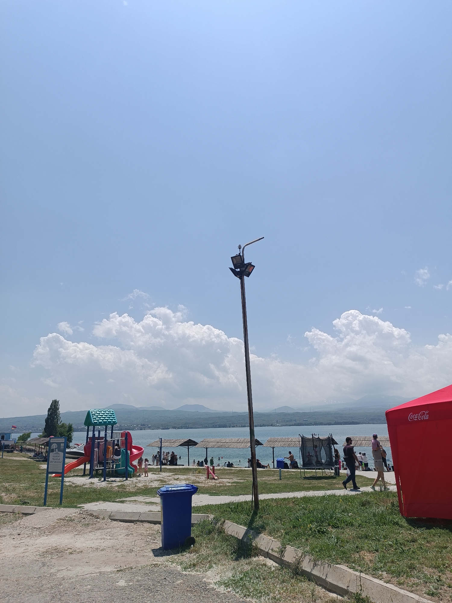A bright, sunny day at a beach in Sevan, Armenia, unfolds with a clear blue sky dotted with white, puffy clouds. In the foreground, a blue trash can sits on a paved area adjacent to a grassy strip. A tall lamppost with multiple lights stands prominently on the left side of the frame, casting no shadows due to the overhead sun.
Further back, a vibrant red Coca-Cola tent is pitched on the right. Beyond this, a sandy beach stretches along the shore of a large body of water, likely Lake Sevan, with a mountainous landscape visible in the distance. Several people are scattered across the beach, some under thatched umbrellas for shade.
To the left, a colorful playground with slides and climbing structures stands on the grass, attracting a few children. A small boat is docked nearby, and a sign with Armenian text is also present. In the middle ground, a trampoline is set up, with a few people appearing to be using it. People are seen walking and relaxing on the beach and along the pathways. The overall scene depicts a lively recreational area on a warm summer day.