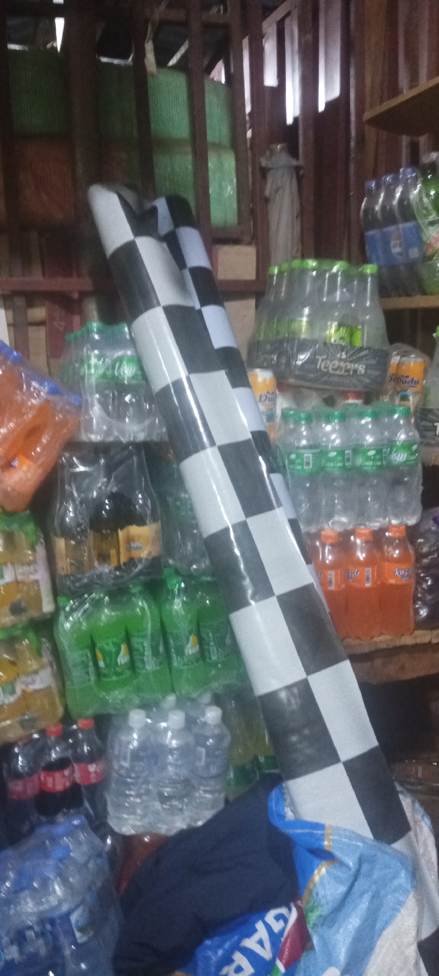The image shows the interior of a small shop in Barakin, Nigeria. Shelves are filled with various drinks, including water bottles, Coca-Cola, Sprite, Fanta, and bottled juice brands. A large roll of black and white checkered material leans against the shelves. A blue bag with text "GAR" is partially visible at the bottom. The scene appears cluttered with the shop's inventory. There are no people visible in the image.