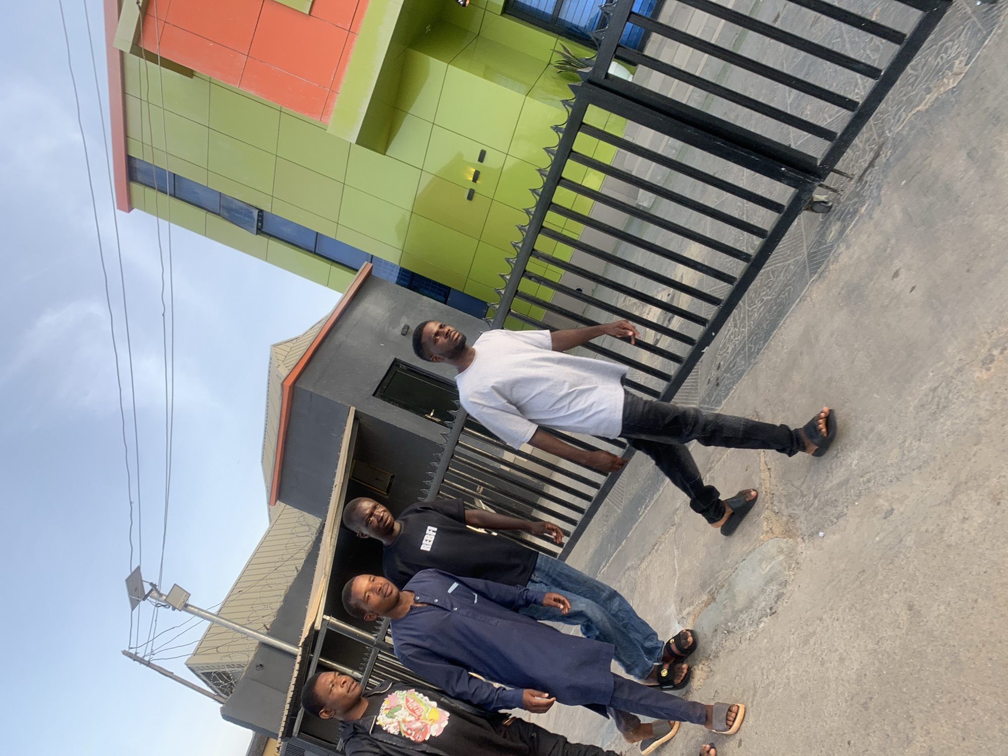 The image captures four young men walking along a sidewalk in the city of Jos, Nigeria. In the foreground, the men are dressed in casual clothing: one wears a navy-blue traditional African tunic, while the others wear jeans and tee-shirts. The man closest to the camera is wearing a black shirt with a floral design. Behind the men is a black metal fence that runs along the edge of the sidewalk. Behind the fence is a building with green and orange paneling. The sky is a pale blue and there are electrical wires running along the top left of the image.