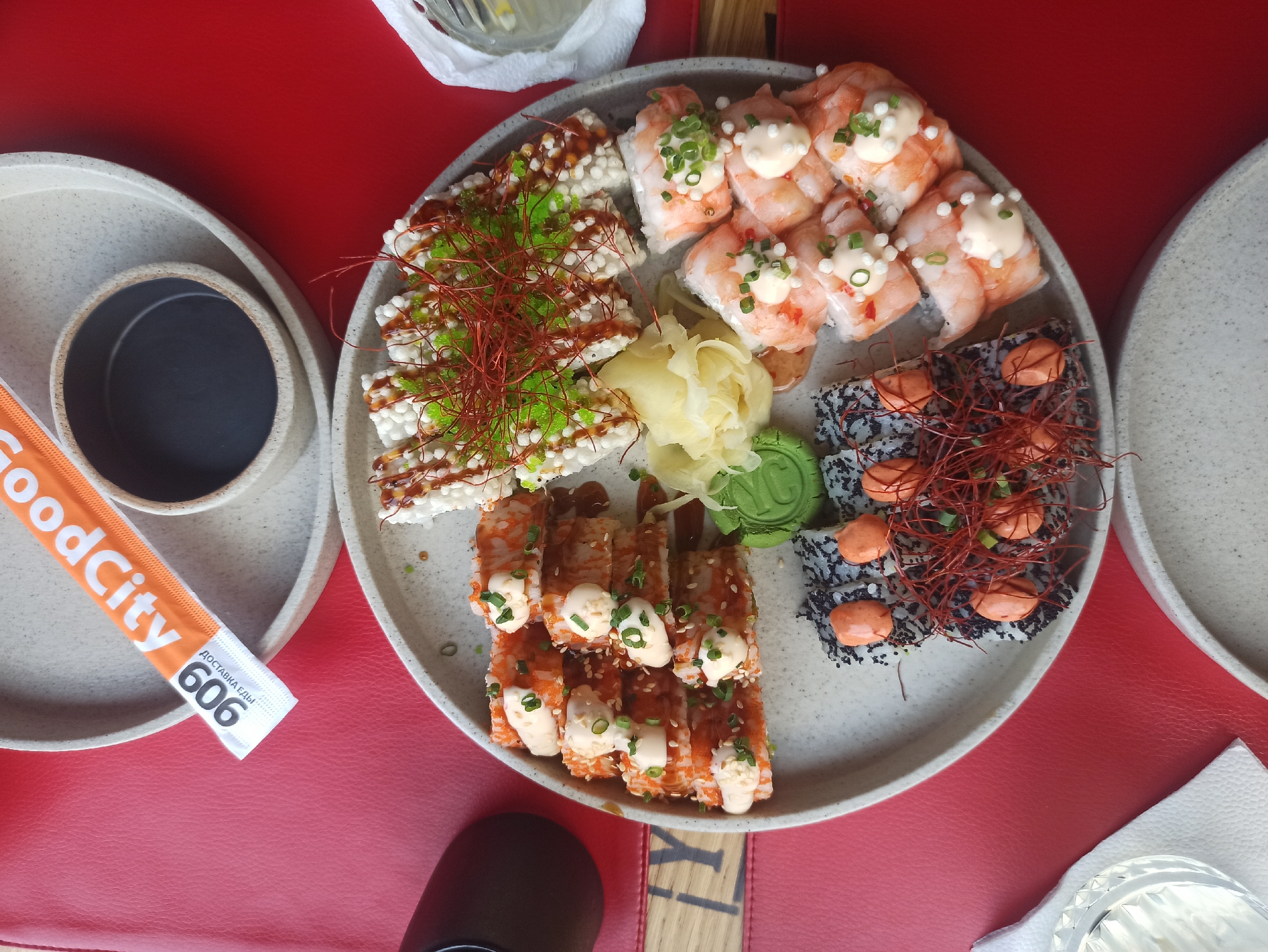 A top-down shot shows a plate of sushi arranged on a red surface, likely a table. The sushi platter contains multiple types of sushi, including shrimp sushi topped with mayonnaise and green onion, sushi with a layer of puffed rice topped with green roe, and sushi rolls decorated with red pepper threads. Pickled ginger and a small, green, circular garnish (possibly wasabi) are placed in the center of the plate. To the left of the plate is a small bowl, presumably containing soy sauce, sitting on a plate. Adjacent to the soy sauce is an orange paper packet with "GoodCity" and "606" printed on it. Other plates, napkins, and a glass are visible around the main sushi platter, indicating a dining setting. The location of the scene is at an unknown location in an unknown city.