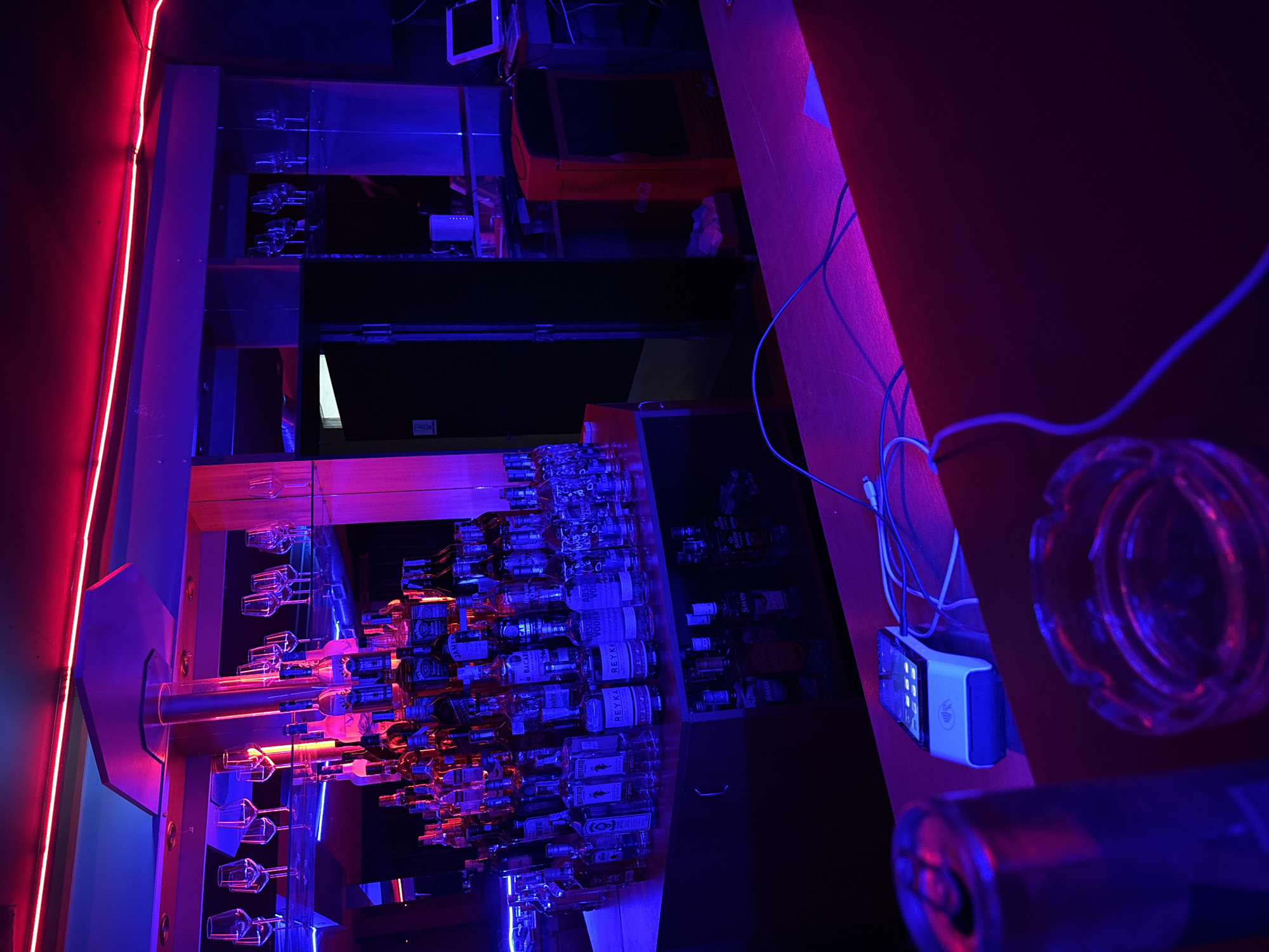 The image depicts the interior of a commercial bar or nightclub establishment. The scene is illuminated by dominant artificial lighting, featuring a red neon strip along the upper perimeter of the room and blue light sources illuminating the bar area.
A central multi-tiered bar structure displays a large assortment of alcoholic beverage bottles, including identifiable brands such as Jack Daniel's, Bacardi, Absolut Vodka, Beefeater, Reyka, Havana Club, Finlandia, and Savoy. Above the bottle display, multiple rows of inverted stemware glasses are racked on transparent shelves. A central vertical support column rises from the bar counter to an overhead fixture.
To the right of the main bar structure, a back bar area features shelves, a red Coca-Cola branded cooler or mini-fridge, and a small electronic display screen (possibly a monitor or laptop) on a counter. A white network device is also visible on a shelf in this section.
In the foreground, on a dark red bar counter, the top portion of a silver beverage can is visible on the left. Adjacent to it, a clear glass ashtray is present. A white electronic payment terminal, connected by cables, rests on the counter. Additional cables are scattered across the counter surface.
No individuals are present in the image, and no specific actions or interactions are depicted. The general ambiance, characterized by the liquor display, specific lighting, and commercial equipment, is consistent with a bar or nightclub environment, located in Varna, Bulgaria, as per contextual information. The precise location within Varna cannot be determined from the visual information provided.