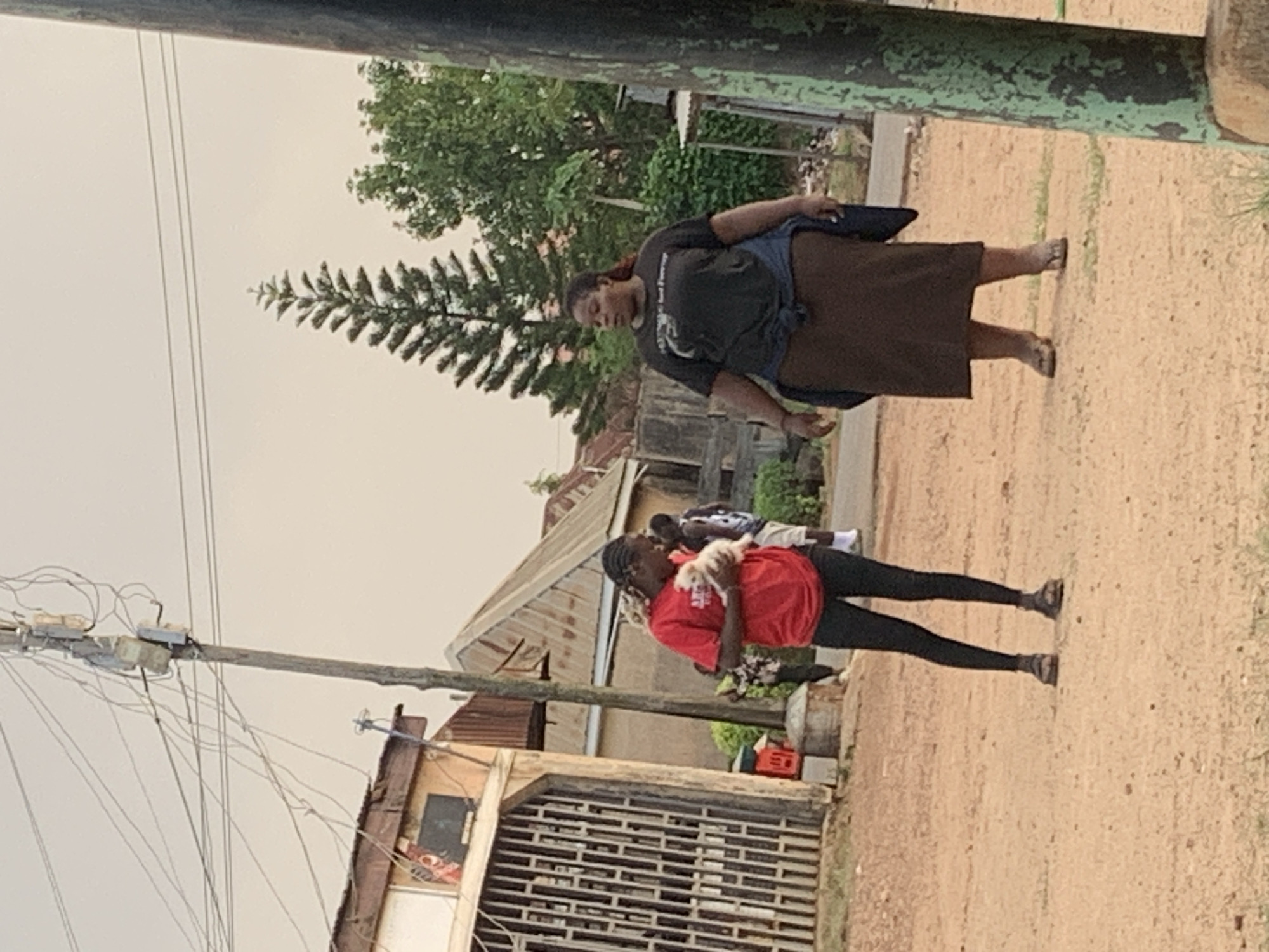 The image, taken outdoors in a residential area, possibly the city of Jos, Nigeria, depicts two women walking on a dirt road. The first woman wears a dark-colored t-shirt, a brown skirt, and sandals. She also carries what appears to be a dark blue jacket or bag over her shoulder. The second woman, who is closer to the camera, wears a red t-shirt, black leggings, and black shoes. She holds a small white dog in her arms. The background includes a corrugated metal fence, buildings with corrugated metal roofs, a utility pole with tangled wires, and trees. The sky is overcast.
