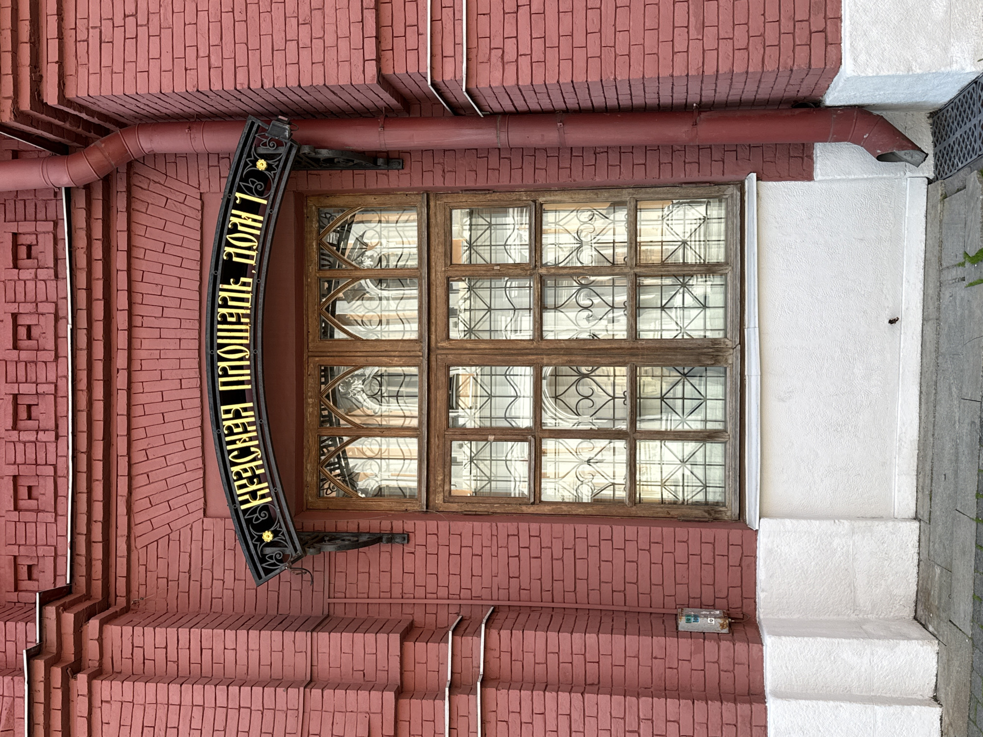 A low-angle, rotated exterior shot showcases a window of a brick building in Moscow, Russia. The building is constructed of red bricks, with a section near the window painted white. A brown gutter pipe runs along the building, above the window. A black sign with gold Cyrillic letters is attached to the building, reading "Красная Площадь Дом 1" (Red Square, House 1). The window is divided into small panes, with some panes having decorative metal inserts. There are steps leading up to the entrance. A small, weathered electrical box is present on the brick wall beneath the window.