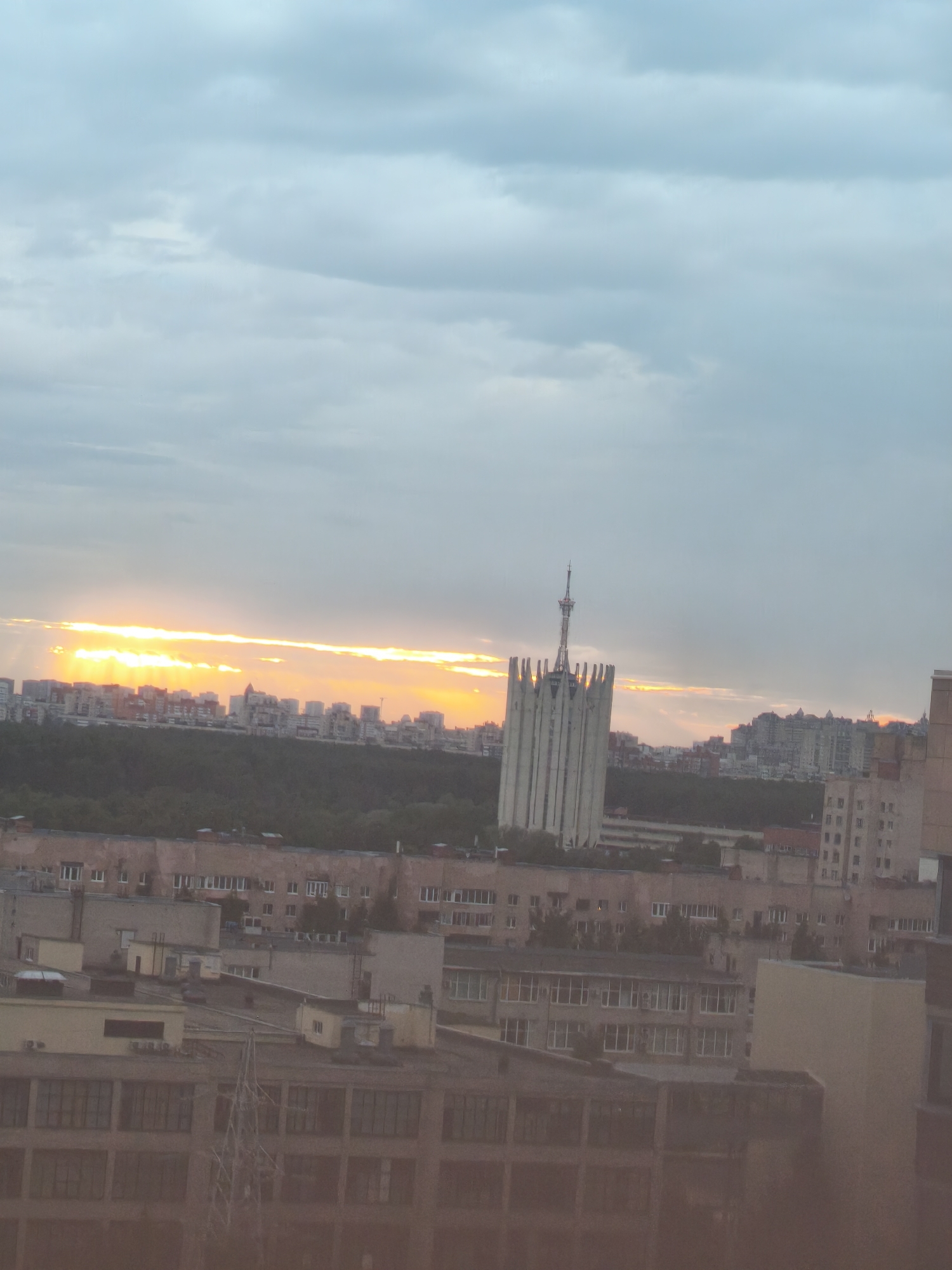 The image presents a cityscape in Saint Petersburg, Russia, taken during a sunset or sunrise. A prominent architectural structure, likely a TV tower or administrative building, is centrally positioned within the frame. In the backdrop, a horizon of buildings is illuminated by the orange and yellow hues of the sun, partially obscured by clouds. Below the horizon line, a densely wooded area contrasts with the urban environment.
The foreground consists of rooftops of residential and commercial buildings, adding layers to the urban landscape. The sky above is filled with a mix of clouds and clear patches, suggesting either an approaching storm or the aftermath of one. The absence of people or vehicular traffic imparts a sense of quietness to the scene.