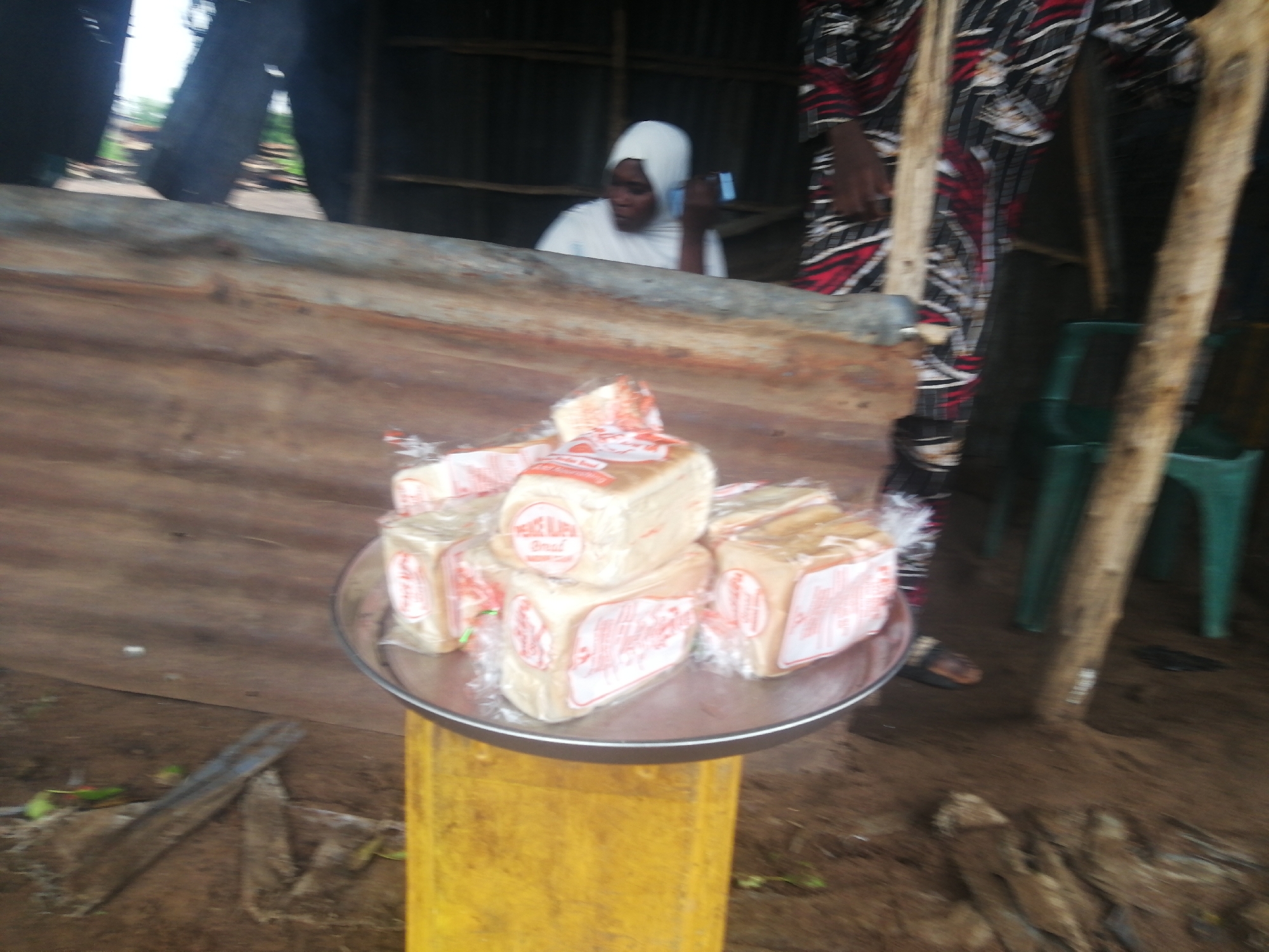 A wide shot captures an outdoor market stall in Omupo, Nigeria. A woman wearing a white headscarf and a white shirt is seen in the background, holding a blue object to her ear, possibly a phone. To her right, a person in a vibrantly patterned African fabric with red, black, and white stripes stands partially visible. In the foreground, a metal tray sits atop a yellow stand, displaying several loaves of bread wrapped in clear plastic with red and white labeling. The stall is constructed from rough wood, with some corrugated metal elements visible. The ground appears to be dirt, and the surrounding environment suggests a rustic, possibly rural, setting within Omupo.
