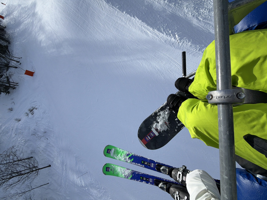 Two individuals are observed from a first-person perspective while ascending a snow-covered mountain slope via a ski lift in Les Allues, France. The foreground displays a grey metal safety bar with a "Sportech" clamp. To the left, a person wearing white pants has a pair of green, blue, and black skis attached to their boots, oriented downwards towards the slope. To the right, another person wearing bright yellow pants has a black snowboard, partially covered in snow, secured to their boots and angled slightly upwards. Below the lift, a vast expanse of white snow is visible, crisscrossed with numerous ski tracks. Sparse, bare trees are present on the left side of the slope, and several orange safety markers are embedded in the snow. The overhead sky is clear and bright blue.
