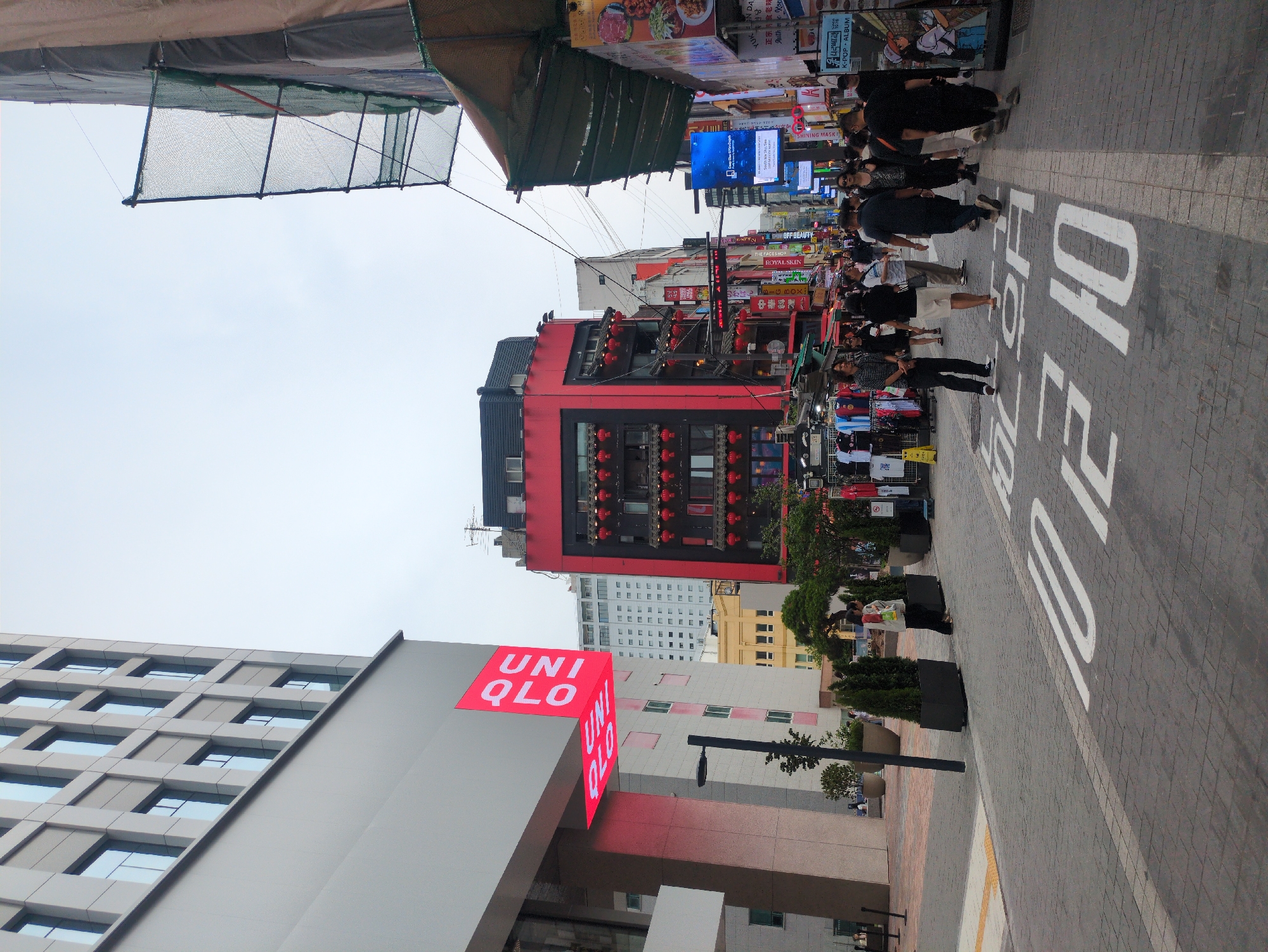 This image captures a bustling urban street scene in Myeongdong, Seoul, South Korea, under an overcast sky.
On the left, a modern, multi-story building with a light grey facade and numerous rectangular windows dominates the foreground. A prominent, illuminated red sign for "UNIQLO" is vertically affixed to its corner, extending outward. Below this building, a paved pedestrian area features rectangular planters with conical evergreen shrubs and a black lamppost.
In the center-right, a distinctive red, multi-story building with a black-tiled roof and traditional East Asian architectural elements is visible. Its facade is adorned with multiple red lanterns hanging from balconies and window frames. This building contrasts with the more modern structures.
To the far right, a building is partially obscured by green safety netting and brown construction tarpaulins, indicative of ongoing construction or renovation. Below this, the street narrows into a commercial thoroughfare lined with various shops, many featuring awnings, Korean and Chinese signage, and outdoor merchandise displays including clothing racks. Large digital advertising screens are visible further down this street.
The street itself is paved with interlocking grey pavers. A wide white line delineates a central path, and large white Korean characters, "천천히" (cheon-cheon-hi), meaning "slowly," are painted on the street surface, indicating a pedestrian-priority zone.
Numerous pedestrians of various ages and genders are walking along the street. Most are casually dressed, with some observing shop displays and others moving purposefully. One individual near the planters on the left wears a light vest and carries a yellow bag. A person further down the street on the left wears a plaid shirt and dark pants, while another in the center wears a dark t-shirt and light trousers. A diverse group of people occupies the central part of the street, with most appearing to be shoppers or tourists. Overhead wires are visible against the sky.