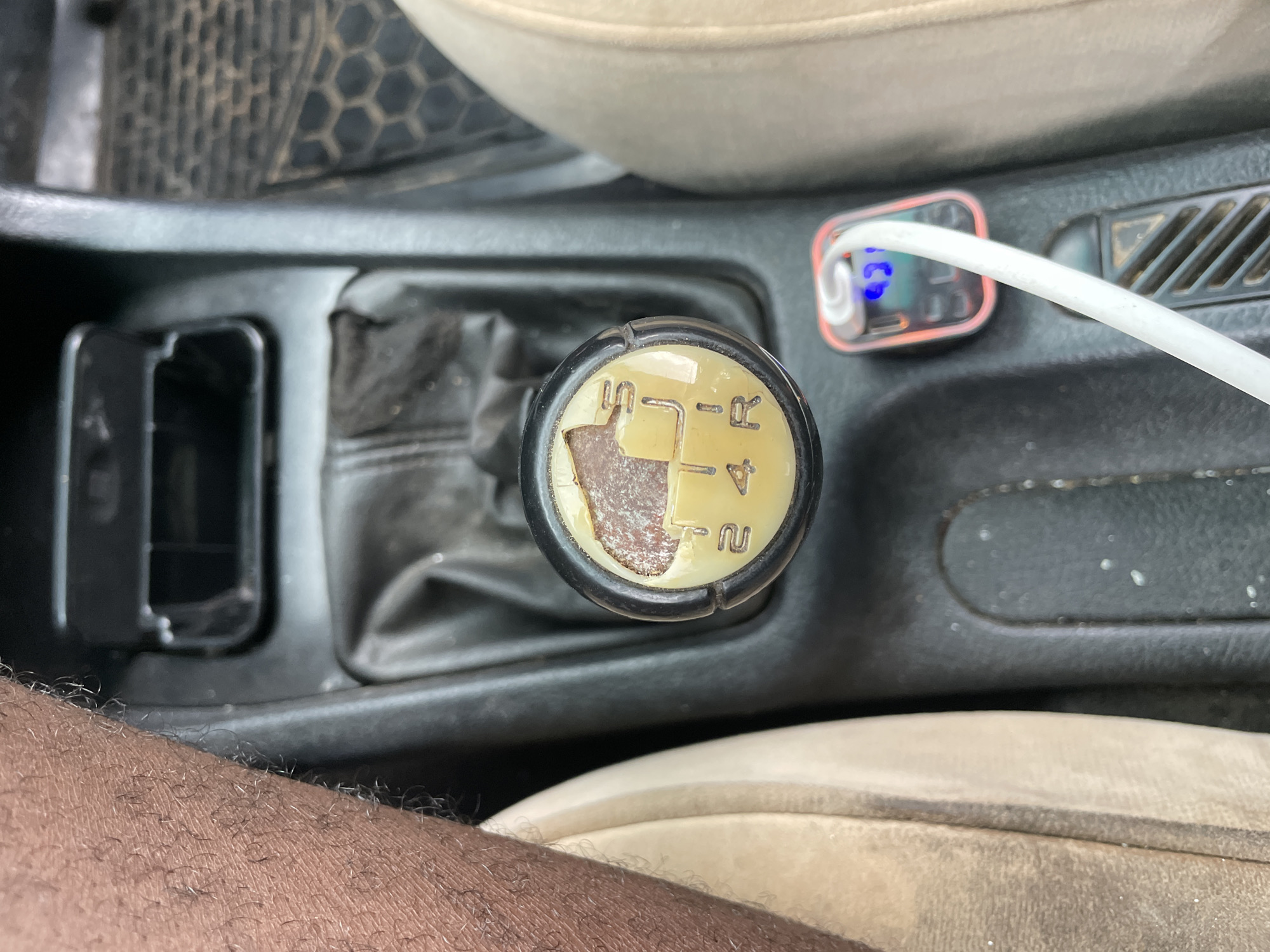 An overhead shot shows the interior of a vehicle, focusing on the gear shift. The gear shift knob displays a standard H pattern, indicating a manual transmission with gears 1 through 4 and reverse (R). The gear shift boot is leather. Beside the gear shift, there is a charging adapter with a USB cable plugged into it. Part of the center console is visible. Also visible are the driver's seat and a portion of the driver's arm. The location is unknown and not definitively specified as Jos, Nigeria.
