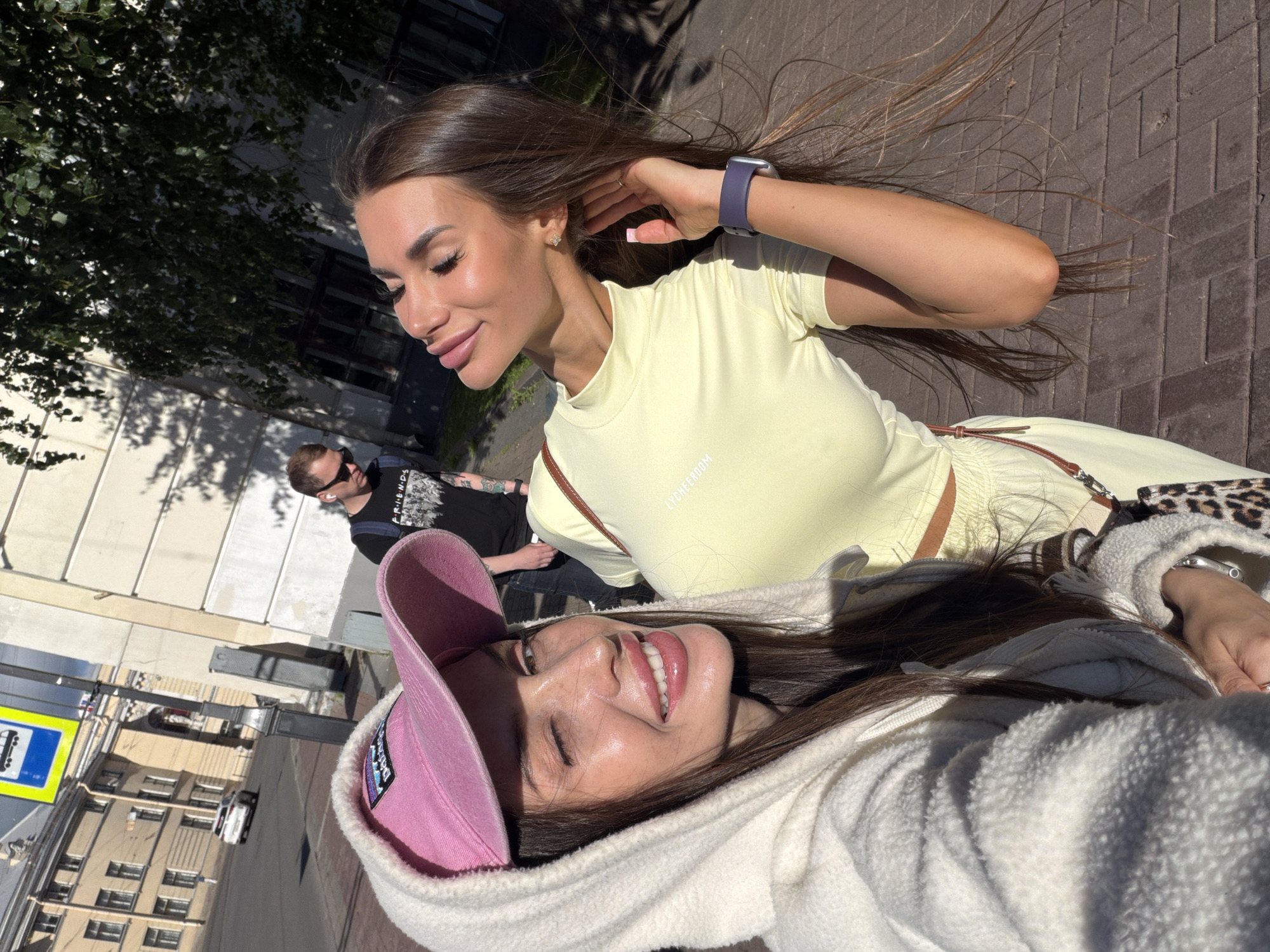 Two women are posing for a selfie on a sidewalk in Saint Petersburg, Russia. The woman on the left is wearing a white hooded jacket and a pink baseball cap. She is winking at the camera. The woman on the right is wearing a light yellow top and has her eyes closed, tilting her head back. She is touching her hair. In the background, a man in sunglasses and a black t-shirt is walking away from the camera. The scene is on a paved sidewalk with buildings and trees visible in the background. The lighting suggests it is daytime and sunny.