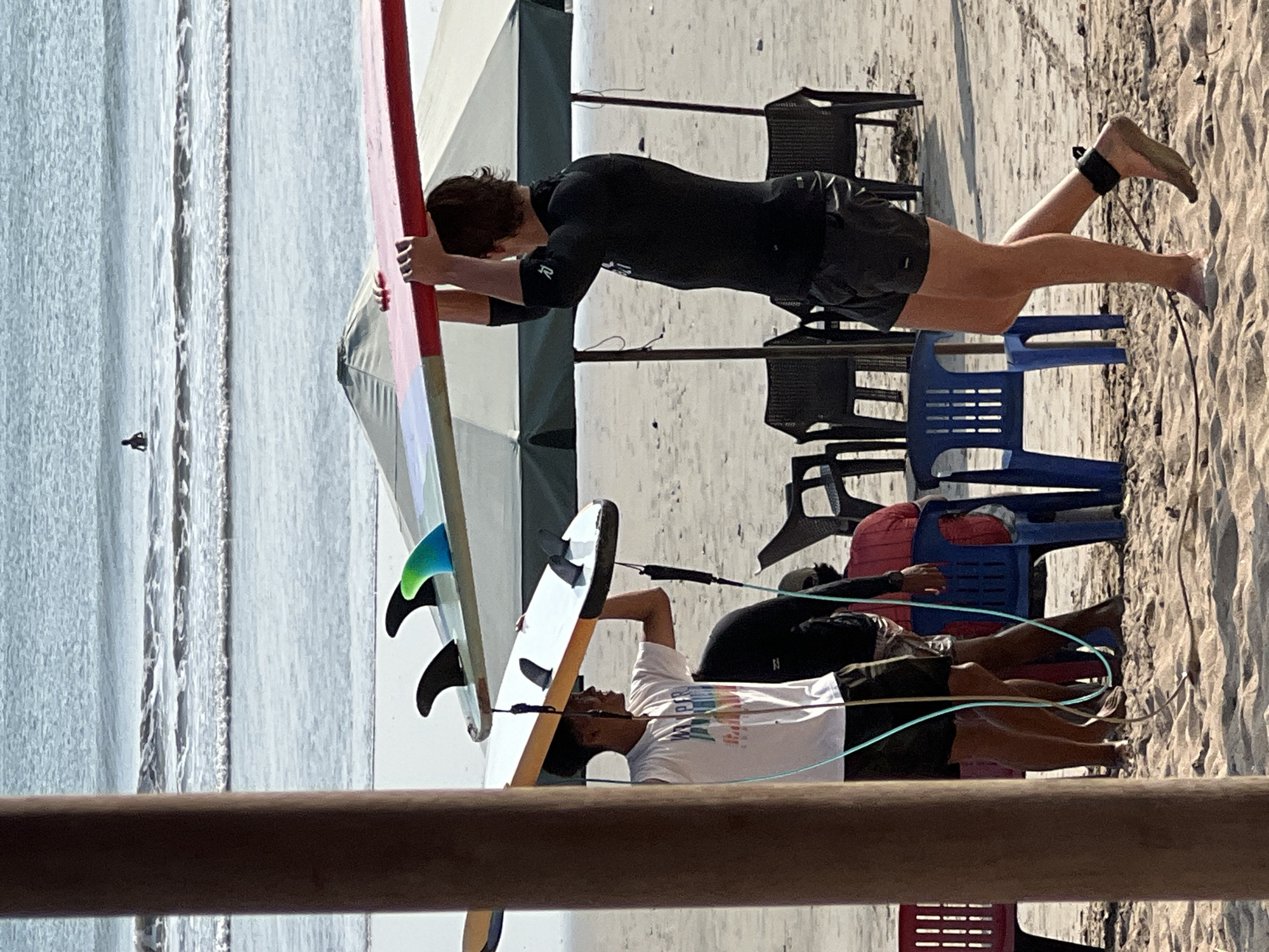 At Kuta Beach, Bali, Indonesia, two young men are carrying surfboards on a sandy beach. The man in the foreground, wearing a black rash guard and black shorts, is walking towards the right, holding a red-topped surfboard above his head. He has a black ankle leash attached to his right ankle. Behind him and to the left, another man in a white t-shirt and dark shorts is carrying a white surfboard with fins up, holding it above his head as well. In the background, a person is visible in the ocean. Beach umbrellas and chairs are set up on the sand. The ocean has gentle waves.