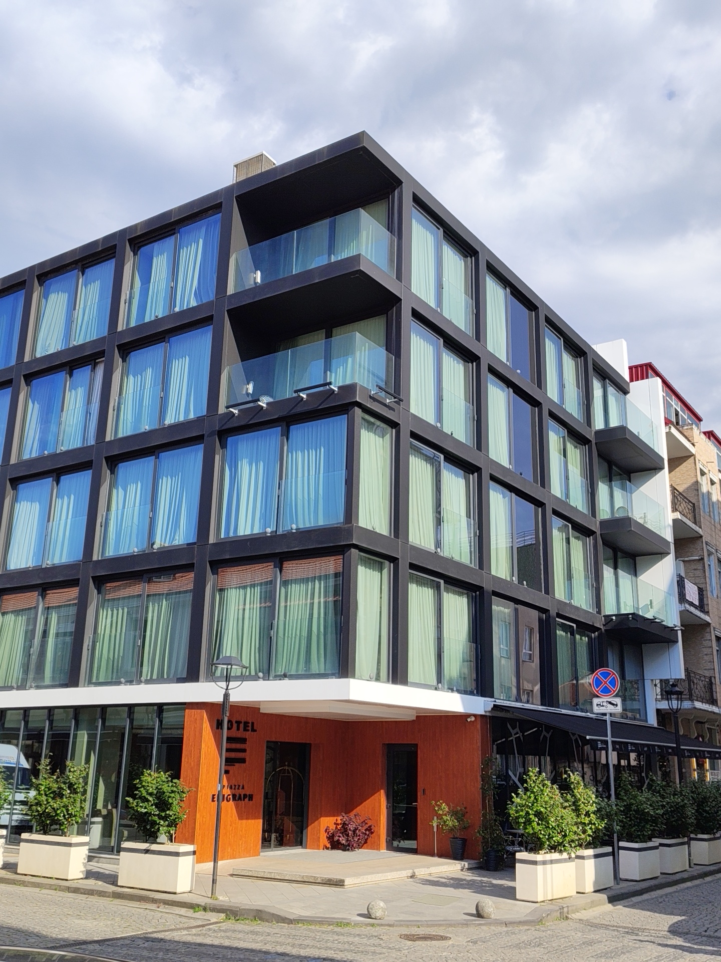The image captures a modern hotel building in Batumi, Georgia. The hotel features a dark-colored exterior with numerous large windows, some with blue tinted glass and others with light green curtains. Glass balconies are visible on multiple floors. The ground floor has an orange-colored section with the word "HOTEL" in black lettering. Planters with green bushes are placed along the sidewalk. In the background, a neighboring building with balconies and a red roof is partially visible. A street lamp and a "no parking" sign are also present. The sky above is cloudy.