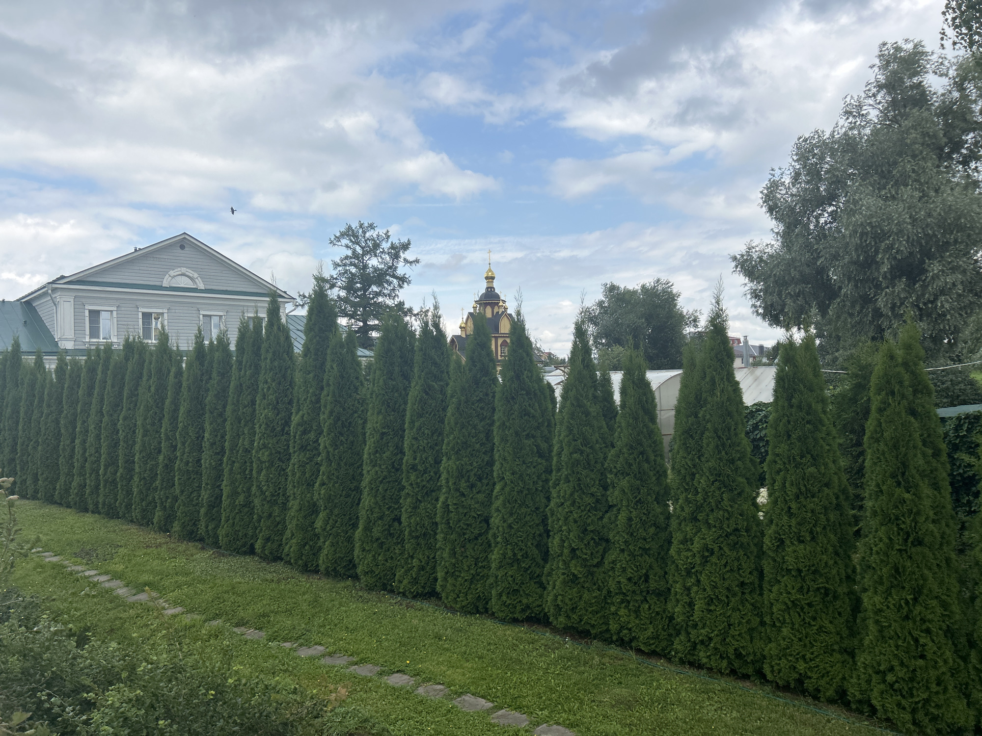 The image shows a serene outdoor scene in Diveyevo, Russia, with a clear view of a meticulously manicured row of evergreen trees forming a natural fence. The trees are densely planted and evenly trimmed, creating a visually striking green wall that extends across the frame. Beyond the trees, a light-colored building with a green roof and multiple windows is visible, suggesting a residential or institutional structure. Further in the background, a prominent golden-domed building, likely a church or monastery, stands out against the sky, characteristic of Russian religious architecture. The sky above is a mix of white clouds and patches of blue, indicating a partly cloudy day. The foreground features a lush green lawn with a stone pathway leading into the distance, adding to the tranquil and picturesque atmosphere of the location. No people are visible in the image.