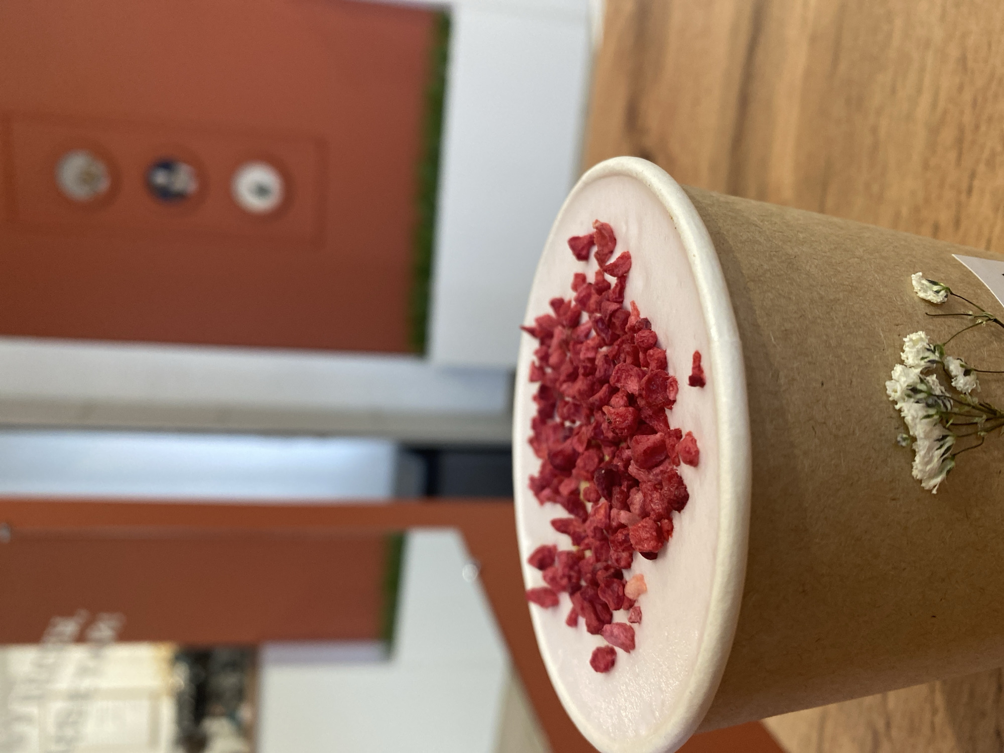 An angled, close-up shot captures a disposable brown paper cup holding a creamy, light pink beverage, generously topped with vibrant red, crushed or freeze-dried raspberries. A small sprig of white dried flowers is tucked into the side of the cup. The cup rests on a light-colored wooden surface, likely a table.
In the blurred background, a reddish-brown wall or panel features three indistinct circular elements, possibly decorative or small fixtures. Adjacent to this, a white wall or surface is visible, with a hint of green vegetation in the far background. The lighting suggests an indoor setting during daytime. The scene evokes a cozy and pleasant atmosphere, typical of a cafe or a relaxed indoor space. No legible text is visible on the cup or in the surroundings. No specific location cues beyond general indoor elements are present in the image.