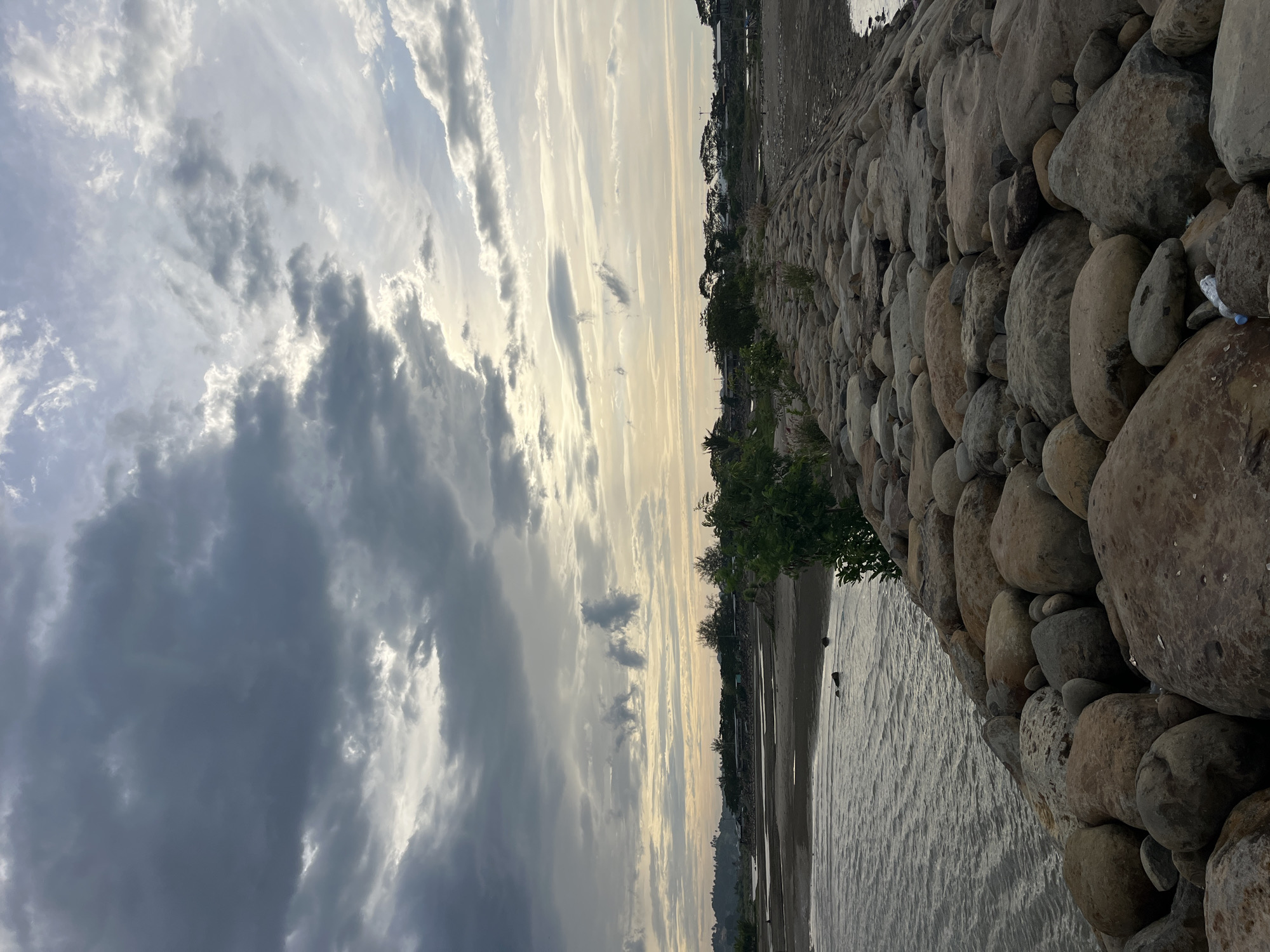 The image shows a view of the coast in Lhokseumawe, Indonesia. A rock wall runs along the right side of the frame, consisting of various-sized, weathered stones. A few small trees and plants grow along the top of the wall. To the left of the rock wall, there's a narrow sandy beach leading to calm water. In the distance, there is a hazy coastline with trees and possibly buildings. The sky is filled with dramatic clouds, ranging from dark gray to light, wispy formations, hinting at a sunset or sunrise. The image is taken at an angle.
