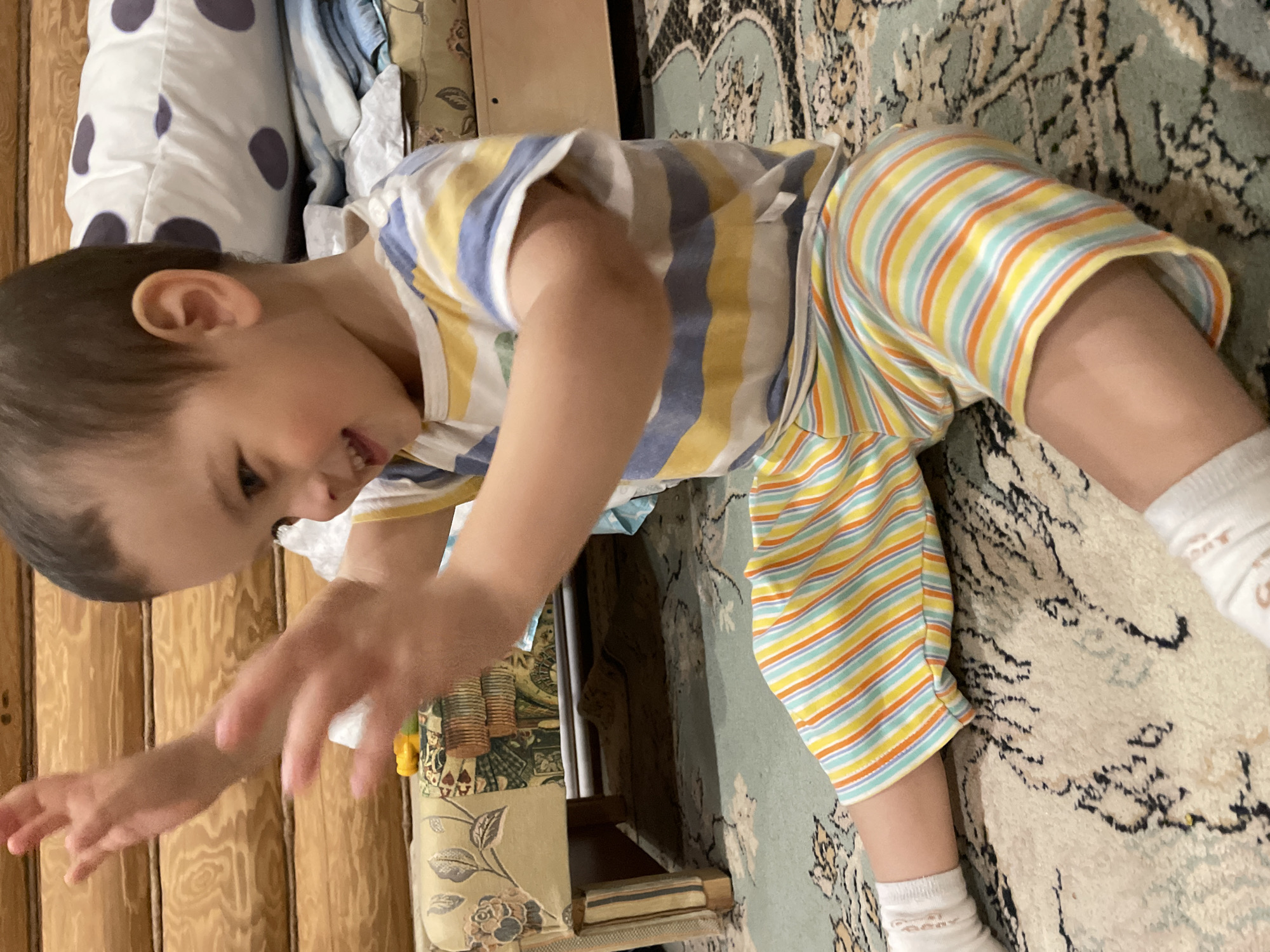 A young child wearing a striped shirt and shorts crawls on a carpeted floor. The background includes a bed with patterned pillows and a wooden wall. The location is unspecified and cannot be determined as Zav'yalovo, Russia.