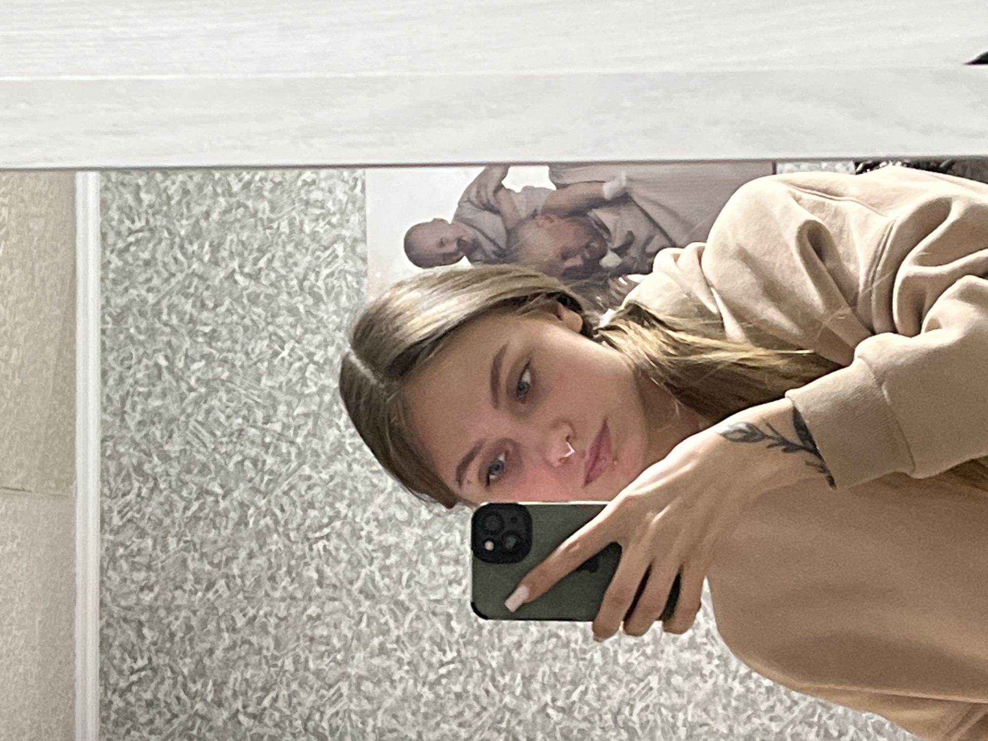 A young woman is taking a selfie in a mirror, inside what appears to be a bathroom with textured wallpaper. The woman is fair-skinned with blonde hair and visible body art on her arm. She is wearing a tan-colored sweatshirt and holding a green iPhone. The location is Фоки, Russia. A picture is also reflected behind her.