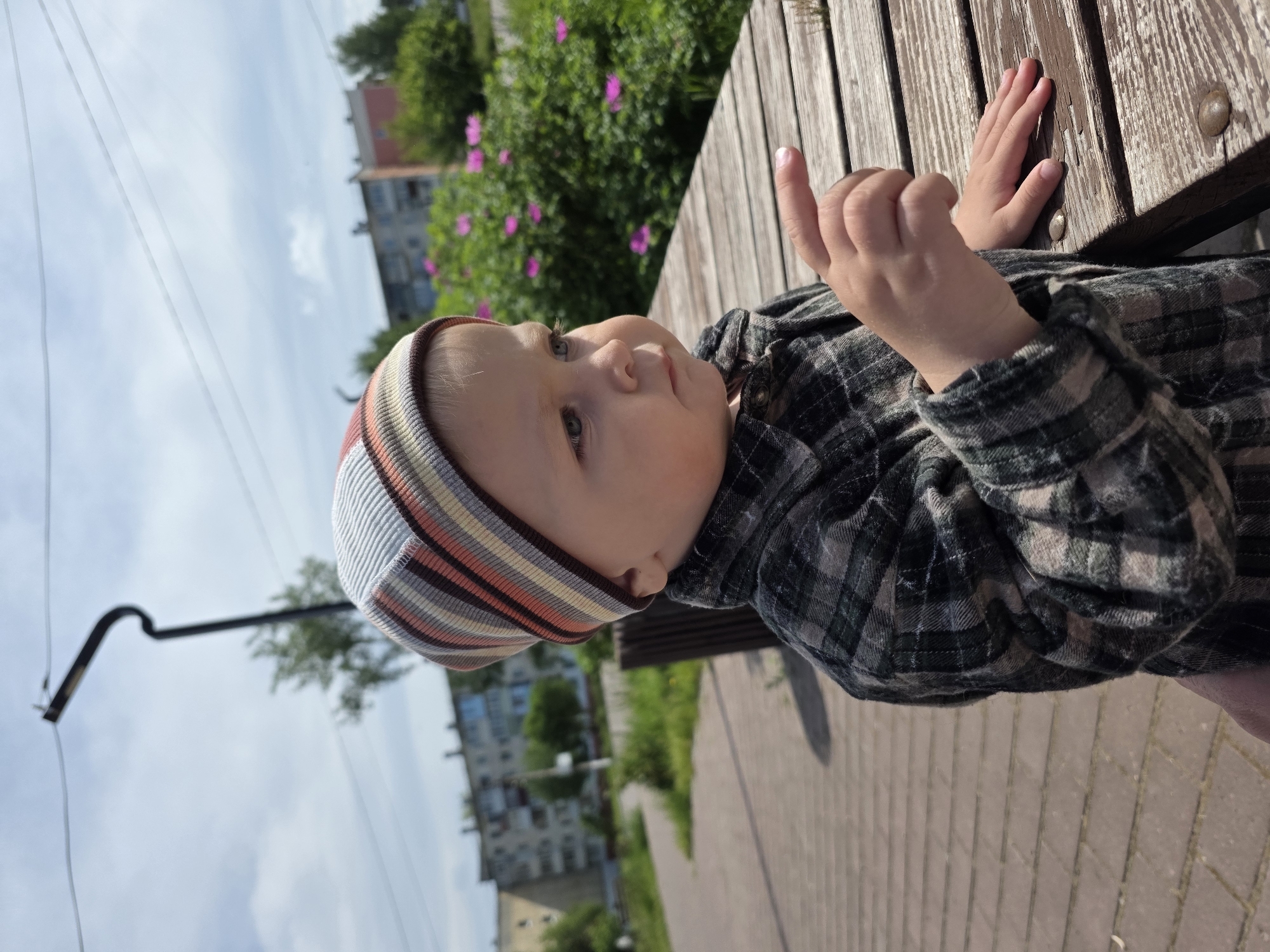 A medium shot captures a young child outdoors, sitting or leaning on a weathered wooden bench. The child wears a multi-striped knit hat in shades of brown, orange, and white, and a dark plaid long-sleeved shirt. Their gaze is directed upwards and slightly to the right, with a neutral, curious expression. The child's left hand is raised in a gestural pose, while their right hand rests on the textured surface of the bench, which shows areas of peeling brown paint and visible metal bolts.
The setting is a public outdoor space, likely a park or walkway in Chaikovsky, Russia. In the mid-ground, green bushes with scattered pink flowers are visible, situated in front of multi-story residential buildings with windows. Overhead, thin power lines stretch across a bright, clear blue sky with some faint white clouds. A black, curved metal pole, possibly a lamppost or utility fixture, is also visible against the sky. The foreground features a paved walkway made of reddish-brown interlocking pavers. The scene is brightly illuminated by natural daylight, indicating daytime. There is no visible text.