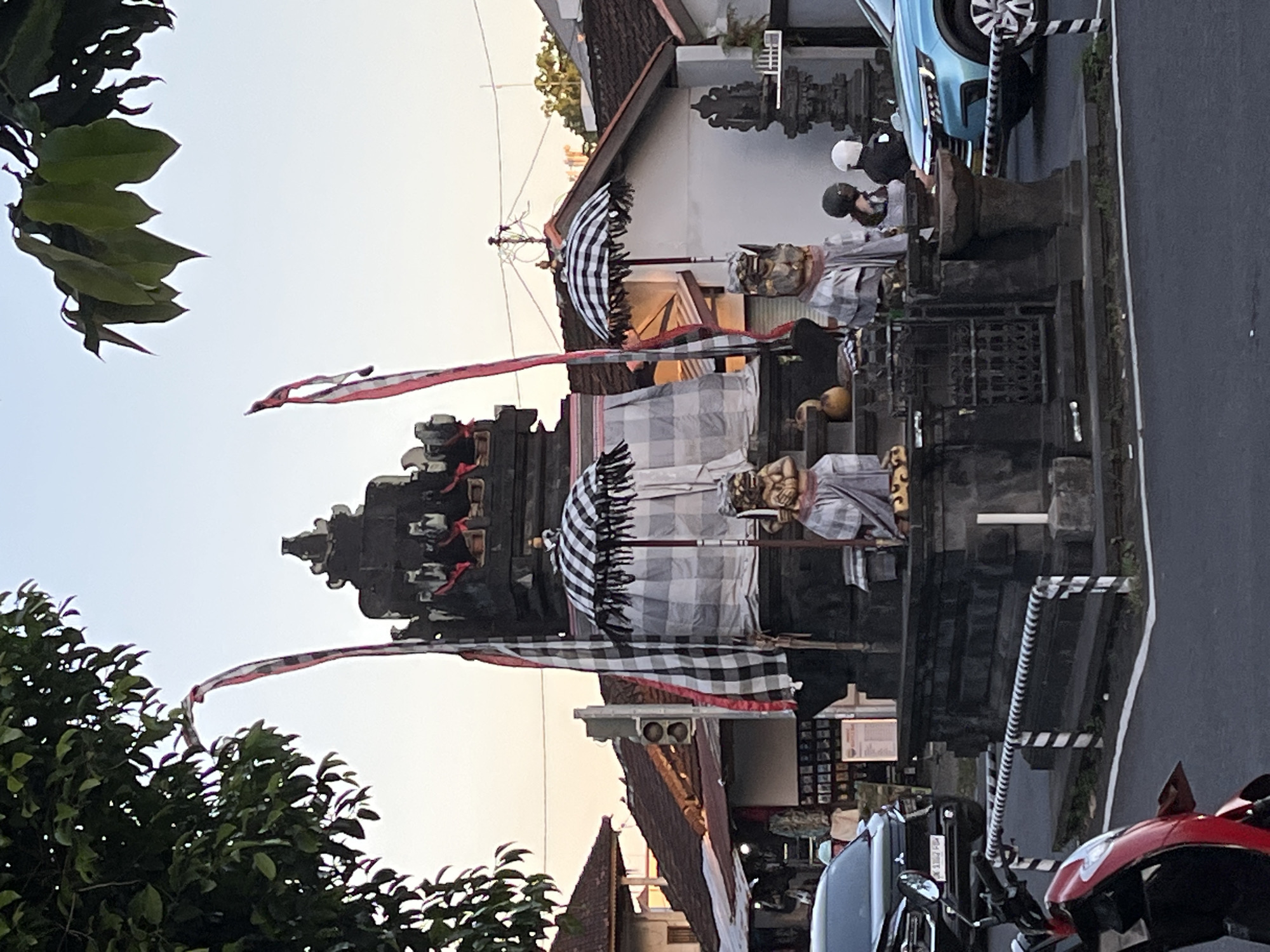The image depicts a temple structure on a street in an urban environment in an unknown city in Indonesia. The temple features stacked, dark gray stone tiers, adorned with a checkered cloth canopy and red and white flags. In front of the temple, there are two statues of figures, draped in checkered cloth. Above the temple, a row of buildings and a parked light blue vehicle can be observed. There are people with helmets on the sidewalk adjacent to the temple. In the foreground, there is a red motorcycle. The photo appears to be taken at dusk.
