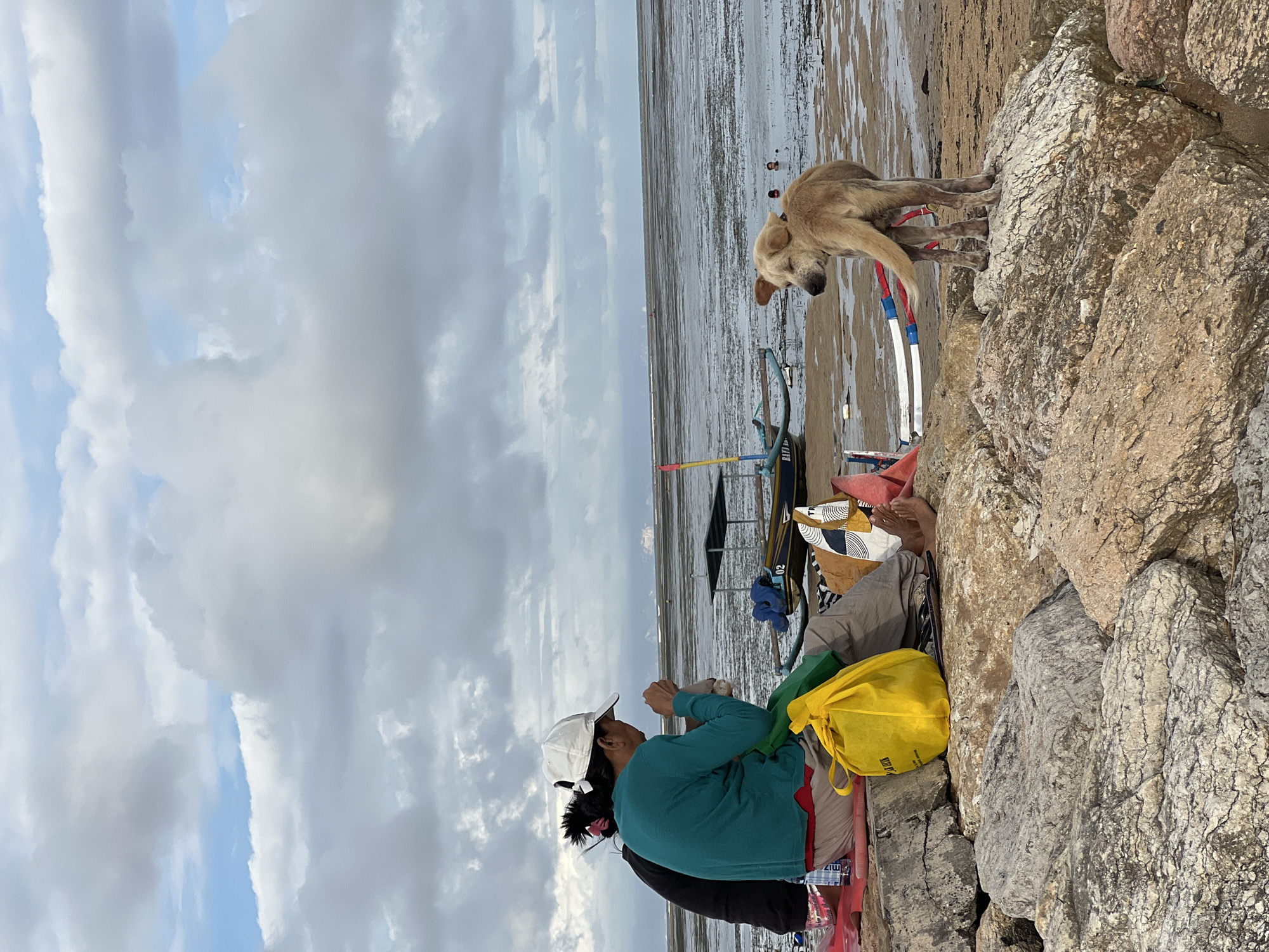 Here is a description of the image:
Two people are sitting on a rocky breakwater. One person is wearing a green long-sleeved shirt and a white cap. The other person is wearing a black top. They are sitting next to a bright yellow bag. Behind them is a sandy beach at low tide. A dog is standing on the rocks near the water's edge. There are several fishing boats near the beach. The sky is overcast with many clouds. The location appears to be a beach in Denpasar, Indonesia.