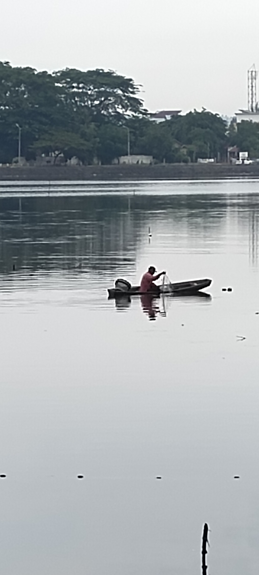 A lone fisherman in a small boat with an outboard motor casts a net on the water. The scene is set in a calm body of water with a reflection of the sky and trees on the surface. In the background, there is a line of trees and some buildings, with a tall industrial structure visible in the distance. The overall atmosphere is serene and quiet. This scene appears to be from the reservoir area in the city of Lhokseumawe, Indonesia.