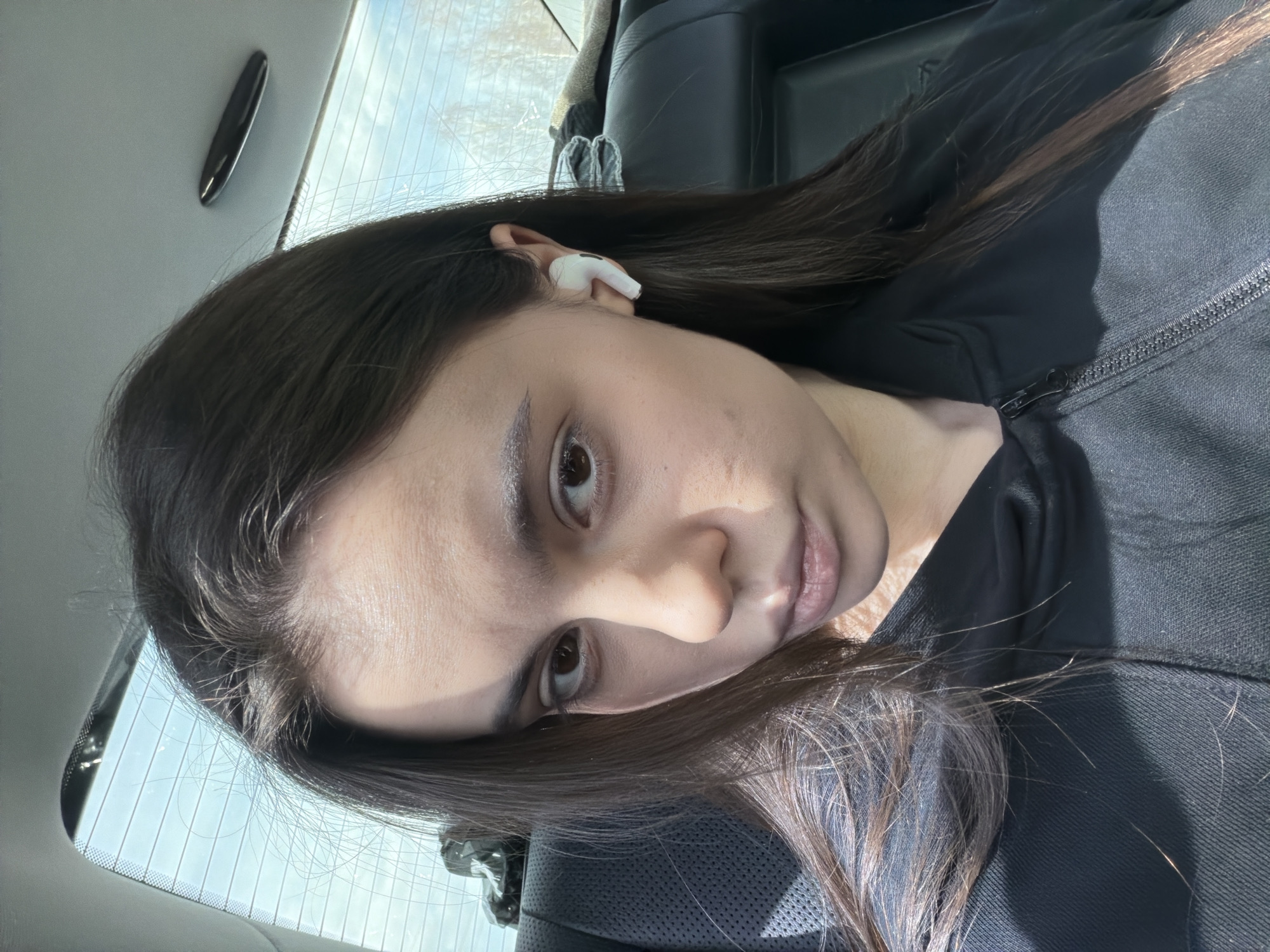 A close-up, tilted portrait captures a young woman inside a car, likely in Ustinovo, Russia, during daytime. She has long, dark brown hair with visible lighter highlights and is wearing a dark, possibly black, zippered top. A white wireless earbud is placed in her right ear. She looks directly towards the viewer with a neutral, slightly serious expression; her lips are slightly parted.
Strong natural light enters the car from behind and to the side, creating distinct shadows across her face and body. The background reveals a car interior with dark upholstery and the rear window, showing clear defroster lines and bright, possibly sunny, outdoor conditions. A small, dark, elongated object is affixed to the car's ceiling or interior trim above her head. No text is visible in the image.