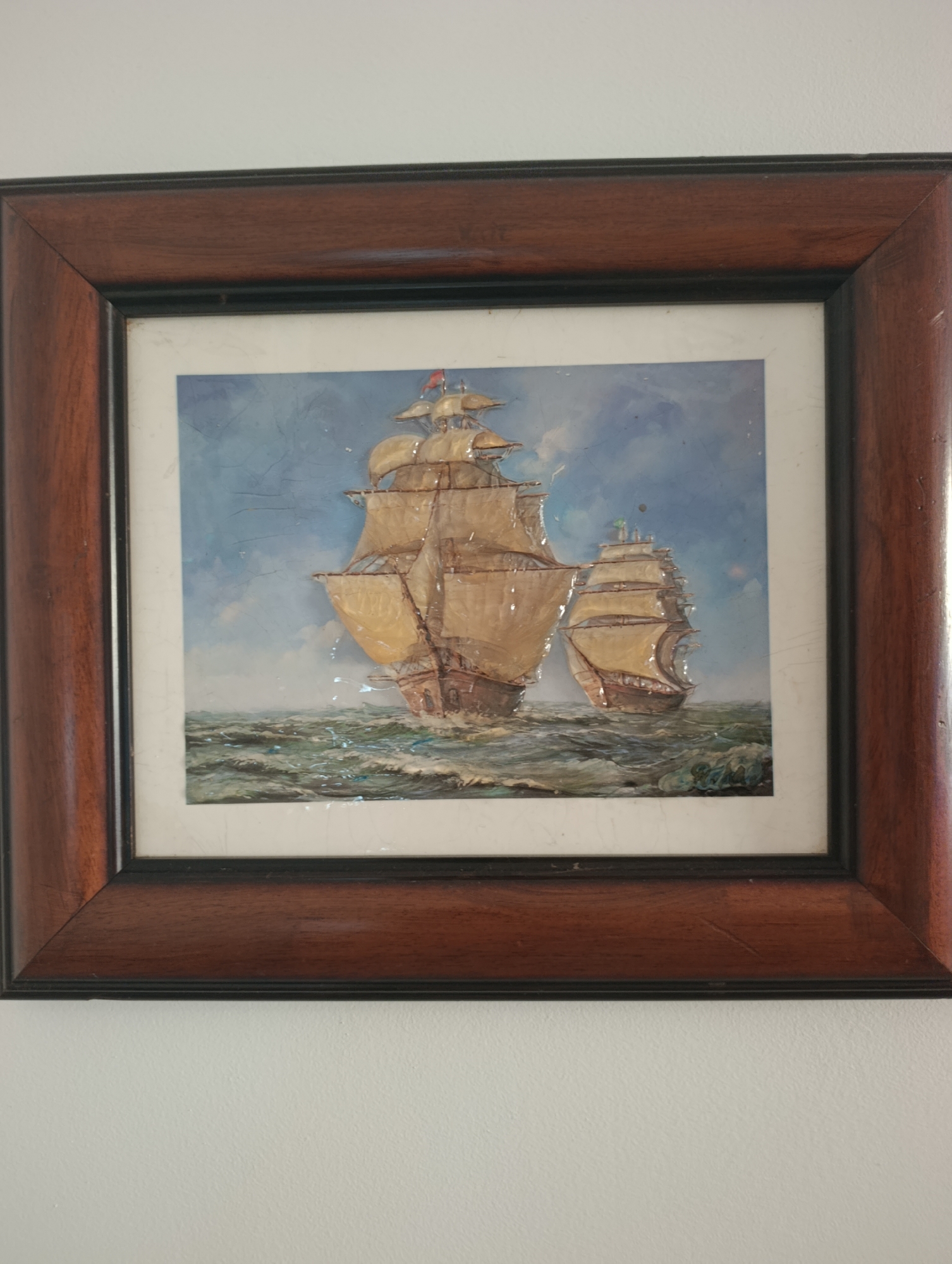 The image shows a framed painting of two tall ships sailing on a choppy sea. The painting is in a dark wooden frame with a subtle sheen. The sky in the painting is blue with some white clouds, and the sea is depicted with white-capped waves. The ships are a light brown color with white sails. One ship is larger and in the foreground, while the other is smaller and in the background. The painting appears to be an oil painting with a textured surface. The painting is hanging on a plain white wall, and the lighting suggests it is indoors. There are no people visible in the painting or in the surrounding environment. The image itself is slightly angled, with the top of the frame closer to the viewer than the bottom. There are some minor imperfections visible on the surface of the painting, such as fine scratches and possibly some glare. The location in Setúbal, Portugal cannot be determined from the image.