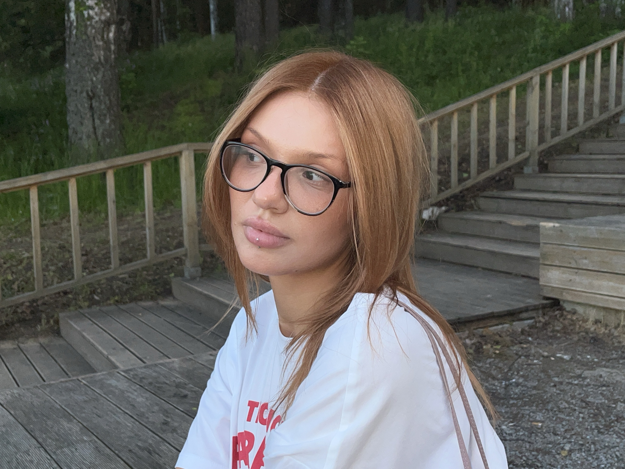 The image depicts a young adult female in Chaikovsky, Russia, captured from the chest up, positioned slightly to the right of the frame, and looking towards the viewer's left. She has long, straight, auburn-colored hair with a distinct central parting. She wears black-framed, circular eyeglasses. A small, silver, spherical labret piercing is visible on her lower lip, positioned slightly to the right of the center. She is dressed in a white t-shirt, with partially visible red lettering on the left side of her chest. The background features an outdoor setting with wooden structures and natural vegetation. To the left of the subject, a light-toned wooden railing runs horizontally, behind which a tree trunk and dense green foliage are visible. To the right of the subject, a set of light-toned wooden stairs ascends diagonally towards the upper right, accompanied by a matching wooden railing. The ground in the lower right foreground consists of dirt and scattered small stones or gravel.