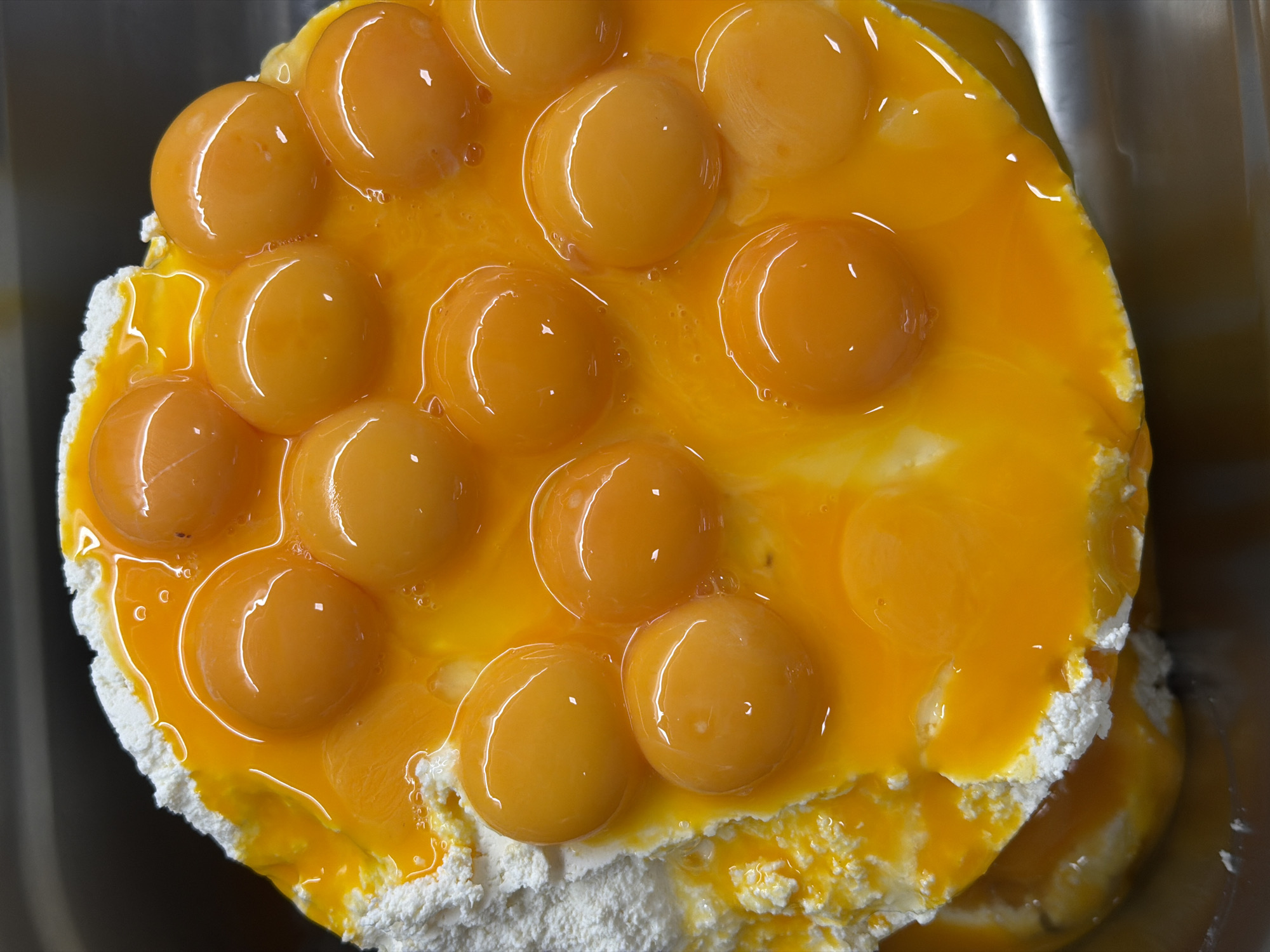 A top-down, close-up shot shows a large quantity of egg yolks atop a layer of white, powdery substance, likely flour or a similar baking ingredient. The yolks, positioned in a somewhat organized, circular pattern, are glistening with a bright orange-yellow color, indicating freshness. Some egg white fluid surrounds the yolks. The scene is captured in a metal container, possibly a commercial mixing bowl. The location and city are not discernible from the image alone.