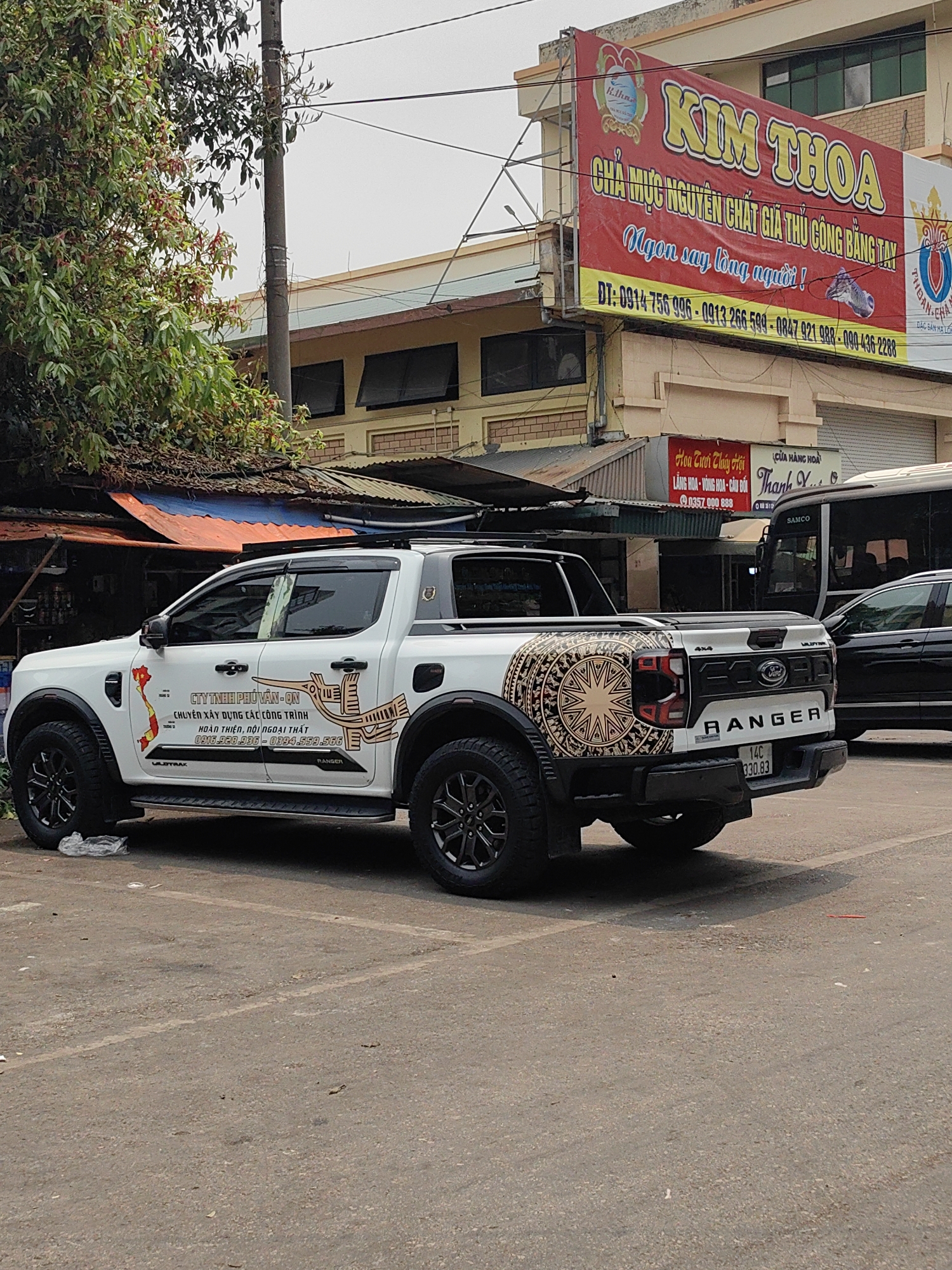 The image shows a white Ford Ranger pickup truck parked on a paved surface in Hong Gai Ward, Vietnam. The truck features unique decorative decals, including a depiction of the Vietnamese map and a stylized bird design, along with a large, circular, patterned graphic on the tailgate. The license plate number is 14C-330.83.
The background includes buildings with Vietnamese signage. One sign prominently displays "KIM THOA" in large yellow letters above a red background, advertising a food product (chả mực). Another sign advertises a flower shop ("Hoa Tươi Thúy Hội").
A section of a black bus or large vehicle is visible to the right of the truck. A tree with green foliage is to the left of the truck. The scene appears to be an urban area with typical Vietnamese businesses and infrastructure.