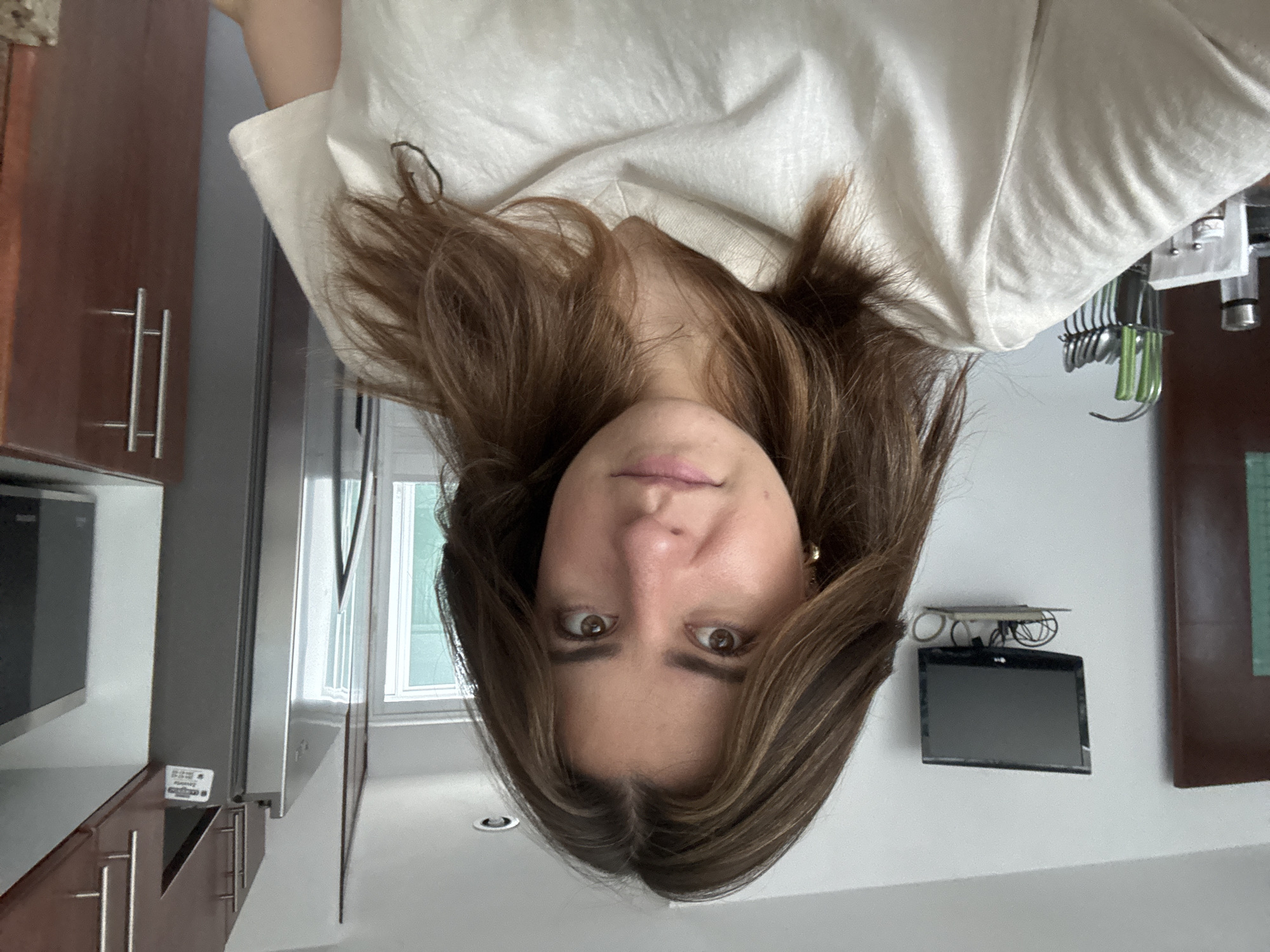 A young woman with long brown hair and brown eyes looks directly into the camera, appearing to be looking down from an overhead perspective. She wears a light-colored (possibly white) top and a gold earring is visible in her right ear. Her expression is neutral, with slightly parted lips.
The background reveals an indoor setting, likely a modern kitchen or an open-concept living space, illuminated by natural daylight coming through a window. To the viewer's left, dark wooden kitchen cabinets with silver bar handles are visible, alongside a stainless steel microwave oven and another stainless steel appliance, possibly a refrigerator or oven. A small white label with barely legible text, including "T-Mobile," is attached near the microwave.
To the viewer's right, a white wall holds a small black screen, likely a wall-mounted television or digital display, with some wires or accessories on a shelf above it. Below this, a rack displays various kitchen utensils, notably a set of cutlery with distinctive bright green handles, alongside other silver utensils. The overall aesthetic suggests a contemporary residential interior. The specific location of Puebla, Mexico, is not visually discernible from the image content.