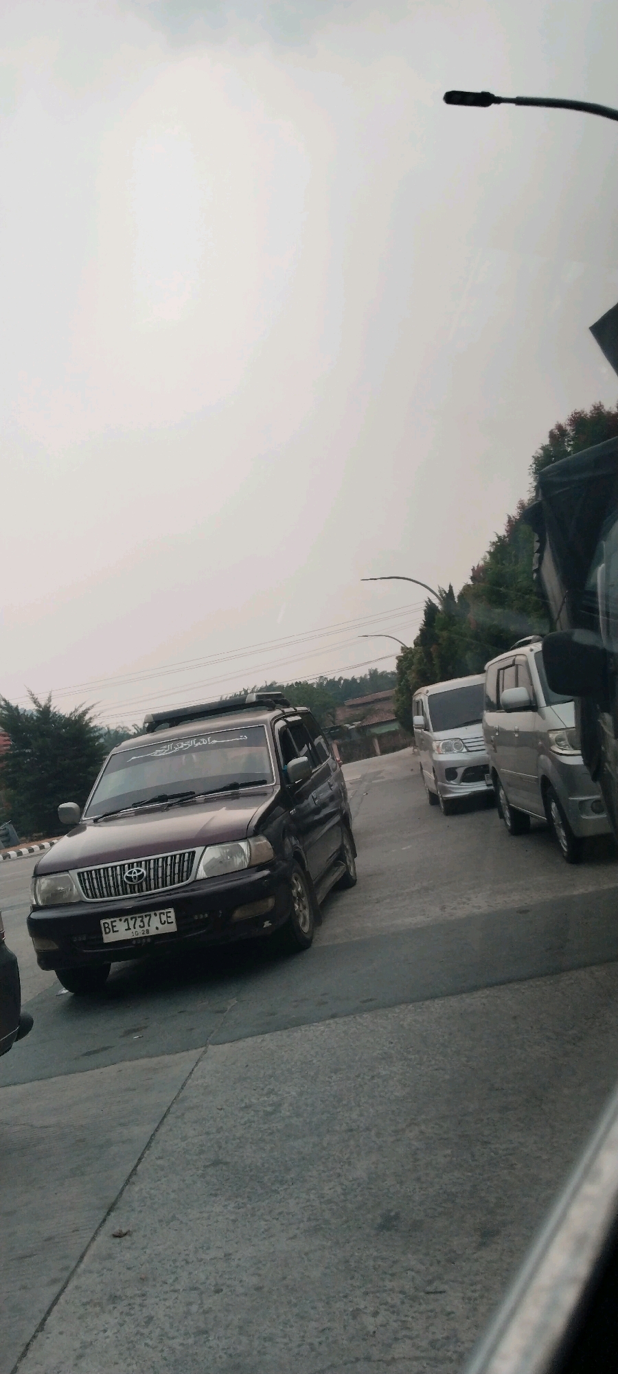 The image captures a street scene in Haduyang, Indonesia, with several vehicles present. A dark-colored SUV, possibly a Toyota Kijang, is prominently positioned in the foreground. It has a license plate that reads "BE 1737 CE". There is text on the windshield of this vehicle. Behind the SUV, a silver van and another similar vehicle are visible, suggesting a traffic flow or parking area. The environment appears to be an outdoor setting with trees and buildings in the background, under an overcast sky. A street lamp is also visible in the upper right portion of the image.