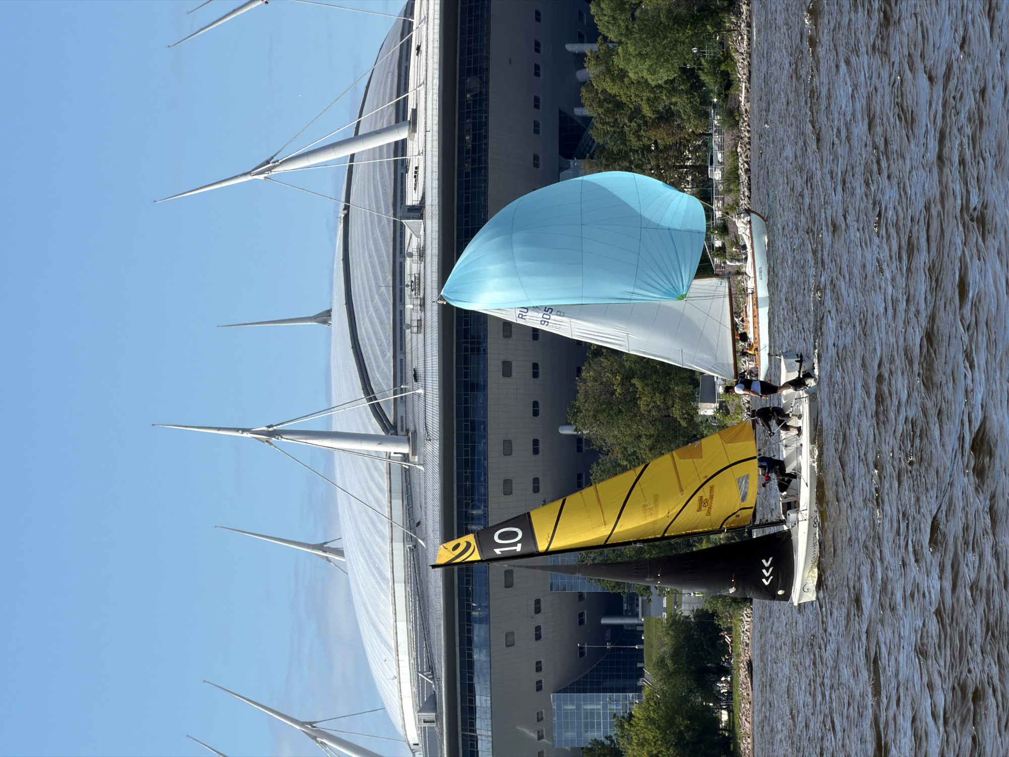 The image depicts a bright, sunny day with a clear blue sky over a body of water where two sailboats are actively racing. In the foreground, a smaller sailboat with a yellow and black sail, prominently displaying the number "10", is positioned to the left. Its sail has black stripes and a black symbol. The boat is crewed by at least two individuals wearing dark clothing. To the right of this boat, a larger sailboat with a light blue spinnaker sail is visible. This sailboat has several people on board, and its sail is marked with "RU 905" in blue lettering. Both sailboats are angled slightly, indicating movement.
In the background, a massive, modern stadium with a domed roof is the dominant architectural feature. The stadium's roof is supported by numerous tall, white, pointed structures. A clear, dark band runs horizontally across the stadium, likely representing a level of seating or a facade. Below this band, a series of rectangular windows are visible. The stadium is situated on the Saint Petersburg shoreline, with a strip of green trees separating the water from the building. Parked cars and other vehicles can be seen behind the trees, suggesting a public area adjacent to the stadium. The water is choppy, with visible waves, indicating a breezy condition. The overall scene captures a sporting event taking place in Saint Petersburg, Russia, with the stadium serving as a recognizable landmark.