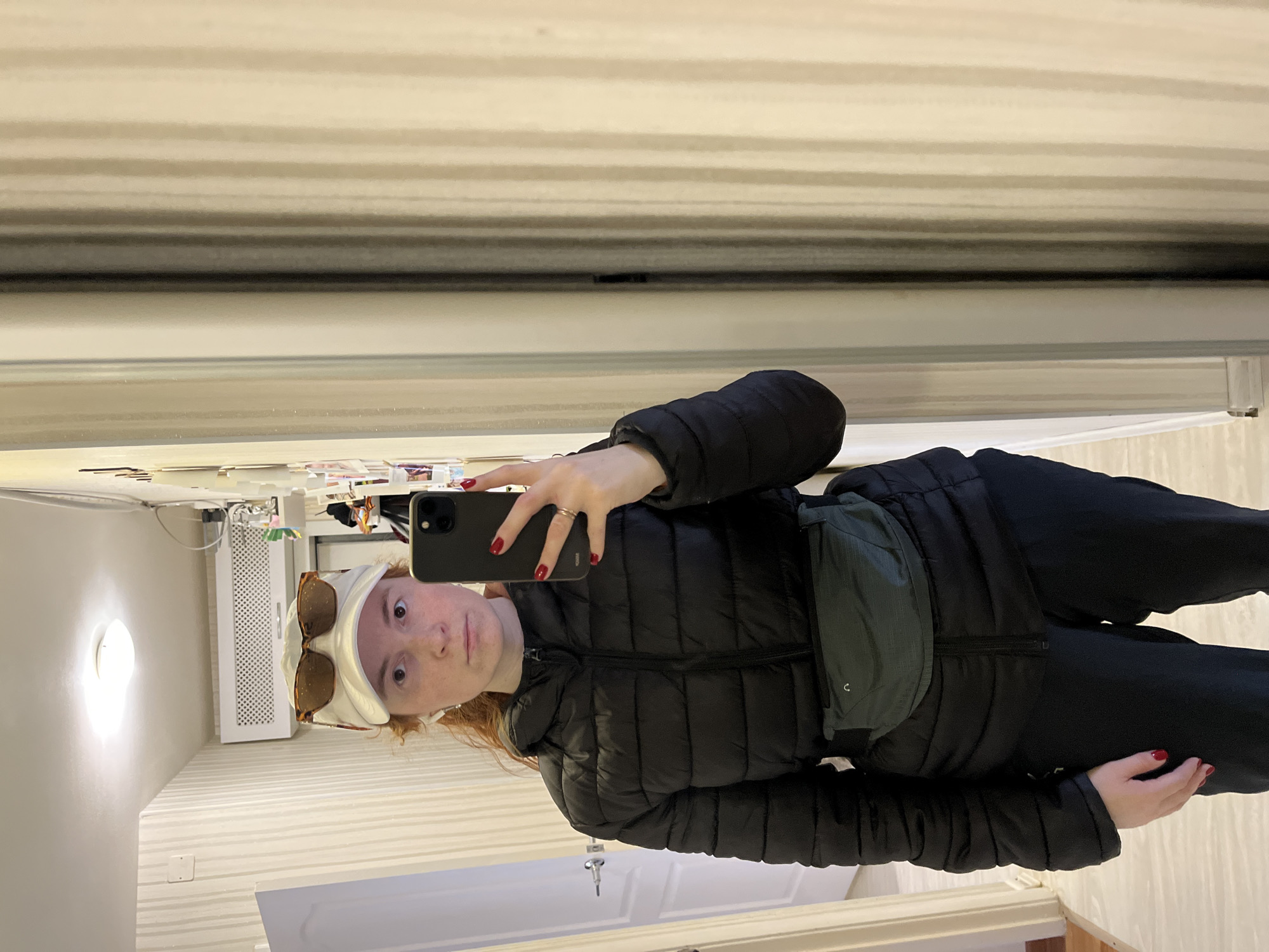 The image, taken in Chaikovsky, Russia, shows a young person taking a mirror selfie indoors. The subject has reddish hair and a neutral expression, looking directly into the camera of a black smartphone held in their right hand. They are wearing a black puffer jacket, dark pants, and a light-colored cap with brown-tinted sunglasses resting on top. A dark green fanny pack or sling bag is worn across their chest. Red nail polish is visible on their fingernails, and a metal ring is on their right hand.
The setting appears to be a well-lit indoor space, possibly a hallway or room. The walls are light-colored and feature a subtle horizontal striped pattern. In the background, to the subject's left, a white radiator or ventilation unit is visible, along with a bright round light fixture on the upper wall or ceiling. Behind the subject's head, on a surface or shelf, various small items or papers are visible, though indistinct. The floor is also light-colored. There is no visible text in the image. The time of day cannot be determined, but the indoor lighting is bright.