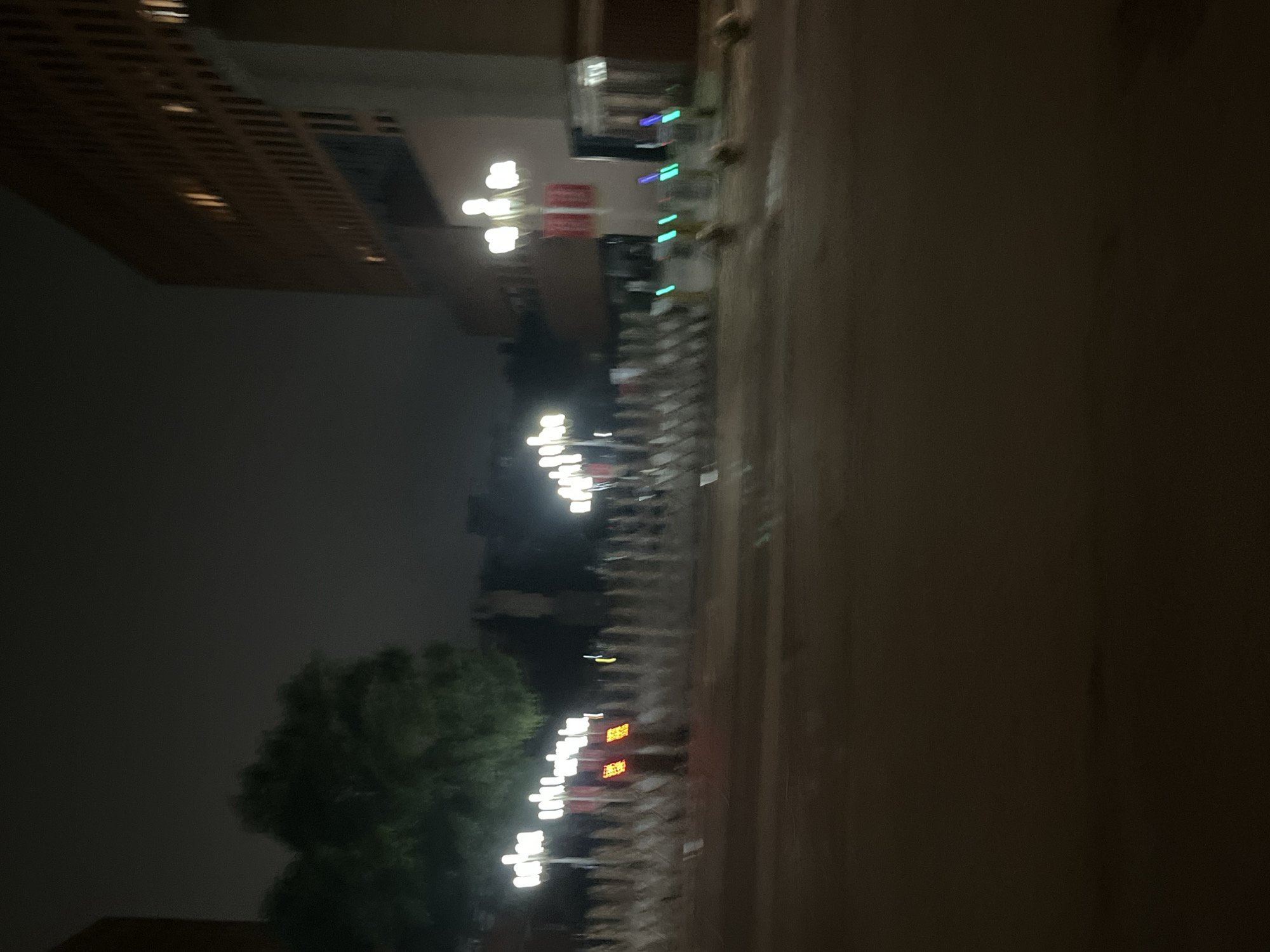 At night, in front of a large building in Yuzhong District, China, a row of white barriers is illuminated by streetlights and other light sources. Two red banners hang from a lamppost, and a traffic light displays red. Several other lampposts are visible, some with multiple lights. In the foreground, the road surface is visible, reflecting some of the light. The overall scene is dimly lit, with the sky appearing dark and overcast.