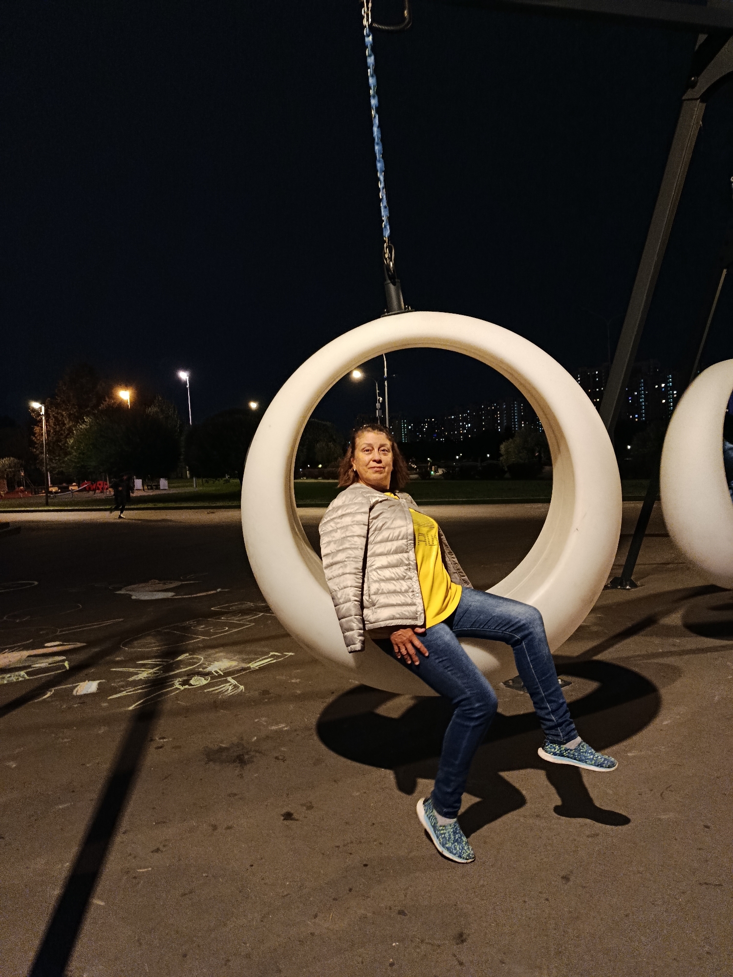 An adult female is seated on a modern, crescent-shaped swing with a white, matte finish. She is positioned within the curved structure, leaning back slightly. She is wearing a yellow t-shirt, a light-colored, quilted jacket, blue jeans, and patterned athletic shoes. The swing is suspended from a dark metal frame, partially obscured by the night sky. In the background, there are streetlights, trees, and distant apartment buildings, suggesting an urban park or playground setting at night. The ground beneath the swing is paved and shows chalk drawings, likely from children. The scene is lit by artificial lights, casting shadows. The location is identified as Moscow, Russia.
