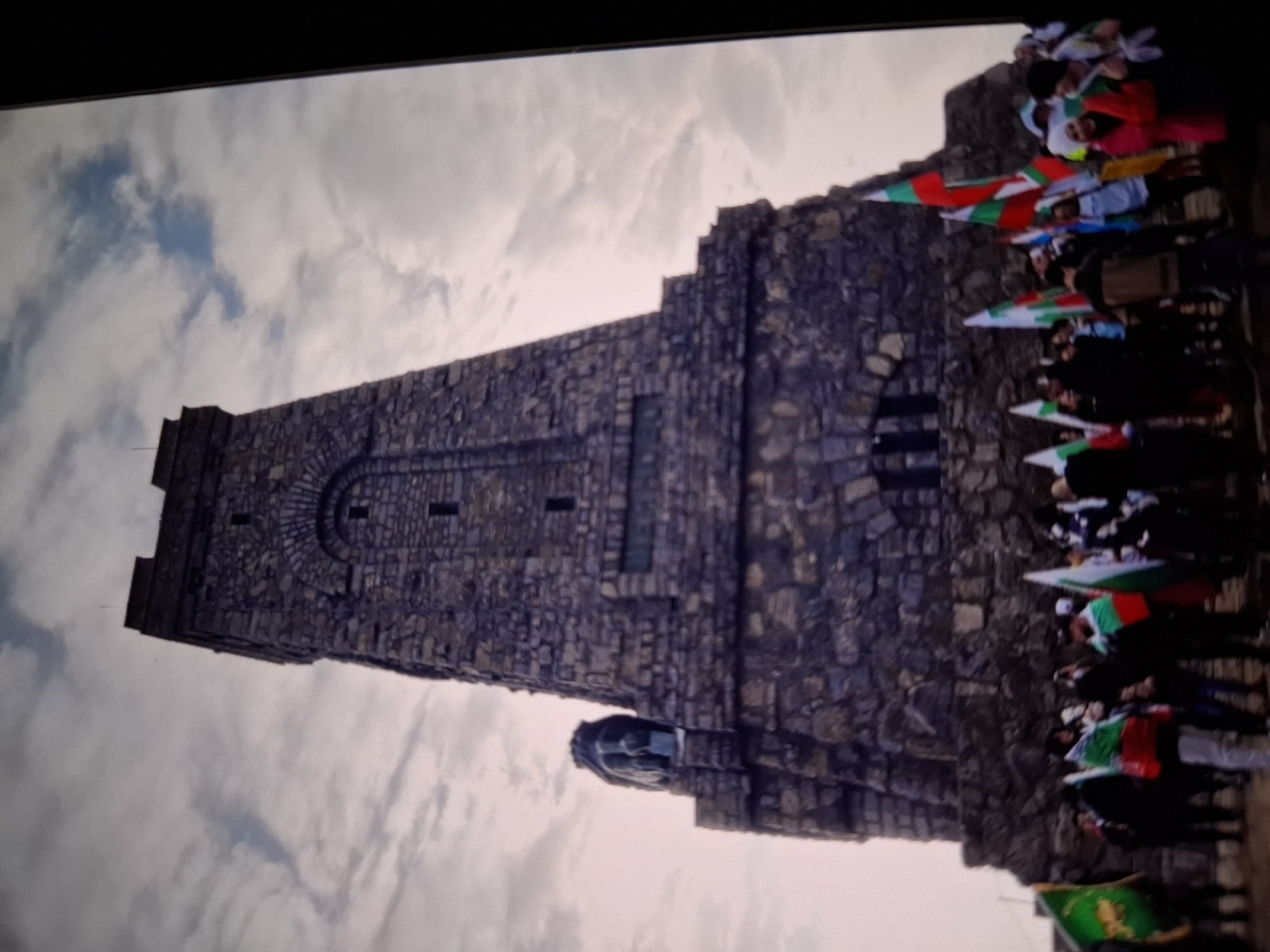 A low-angle, wide shot captures the monument to the defenders of Shipka Pass, a weathered stone tower structure resembling a medieval fortress with multiple levels and arched windows. A large stone lion sculpture is visible on the left side of the monument's base. In the foreground, a crowd of people is gathered, many holding Bulgarian flags, which are white, green, and red. The sky above is overcast with wispy clouds and patches of blue. The ground appears to be dirt or gravel.