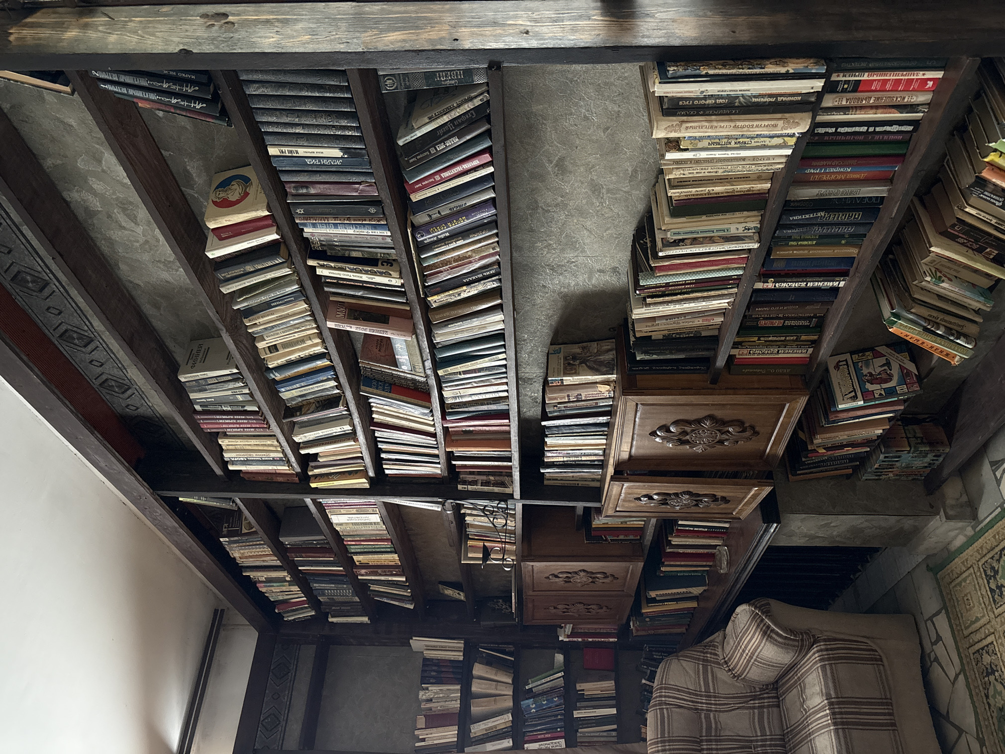 The image shows a corner of a room with a large, dark wooden bookshelf filled with books. The bookshelf is made of thick, rough-hewn wood and extends from floor to ceiling. The books on the shelves are of various sizes and colors, with some spines showing text in Cyrillic script. On the lowest visible shelves, there are also two carved wooden cabinets. To the left of the bookshelf, a portion of a plaid armchair is visible. The room appears to be dimly lit, creating a cozy and somewhat antique atmosphere. The wall behind the books is textured, resembling concrete or stone. The floor is tiled with a geometric pattern, and a patterned rug lies on it. The overall scene suggests a personal library or study, possibly in a home. There are no people visible in the image. The location is identified as Zavod Mikhailovskii, Russia.