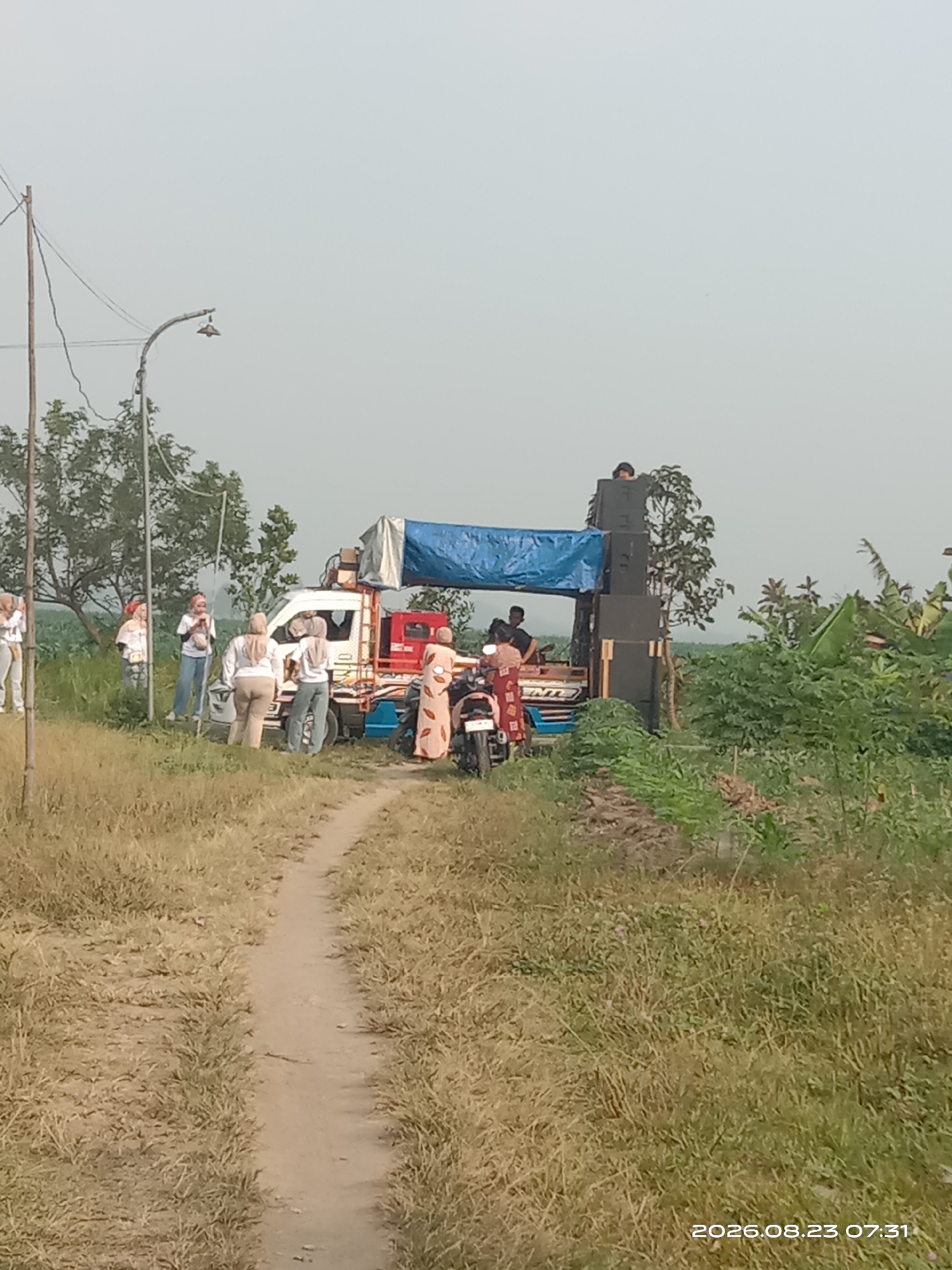 In Banyuarang, Indonesia, on a dirt path surrounded by dry grass and green vegetation, a group of people is gathered around a small truck modified with a sound system. The truck has a blue tarp covering part of its cargo area, and large speaker boxes are stacked on top. Several individuals, mostly women wearing headscarves and casual clothing, are standing near the truck, some appearing to be interacting or posing for photos. A motorcycle is parked to the right of the truck. In the background, trees and a utility pole with wires are visible against a hazy sky. The scene appears to be outdoors in a rural or semi-rural setting.