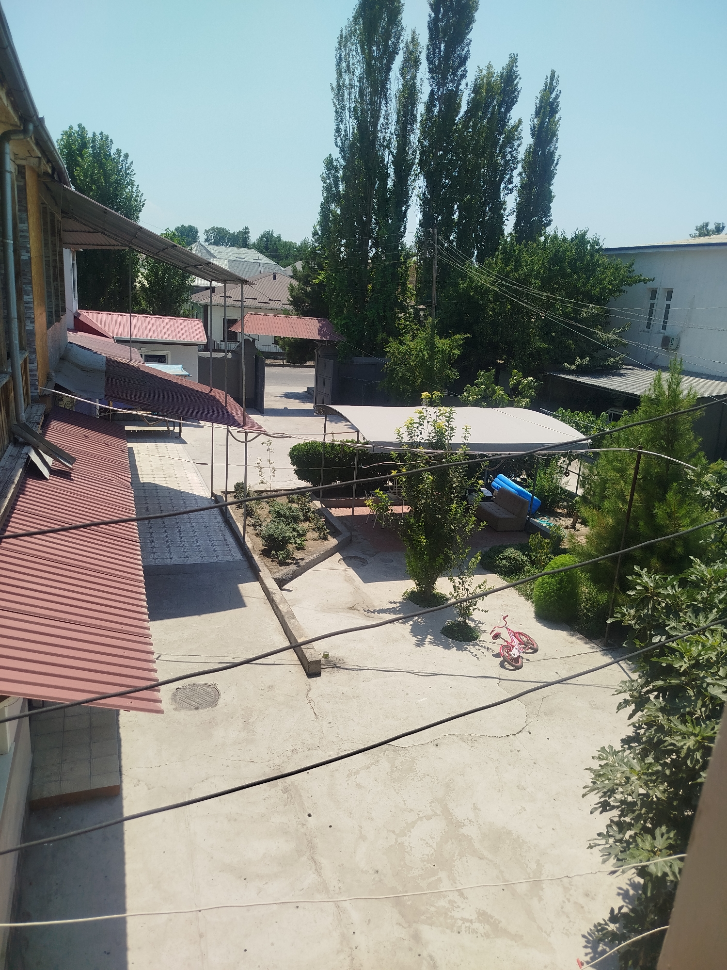 The image depicts a sunny courtyard in Kokand City, Uzbekistan. In the foreground, a concrete ground is partially covered by the shadow of a corrugated red metal roof on the left. The courtyard contains a small garden bed with green plants and a pink children's bicycle lying on its side. Further back, a white canopy shades a seating area with chairs. Beyond this, there's a blue inflatable object and some brown furniture. The background features more residential buildings with red roofs, tall trees, and a white building with windows. Power lines are visible crisscrossing the scene. The sky is clear and blue.