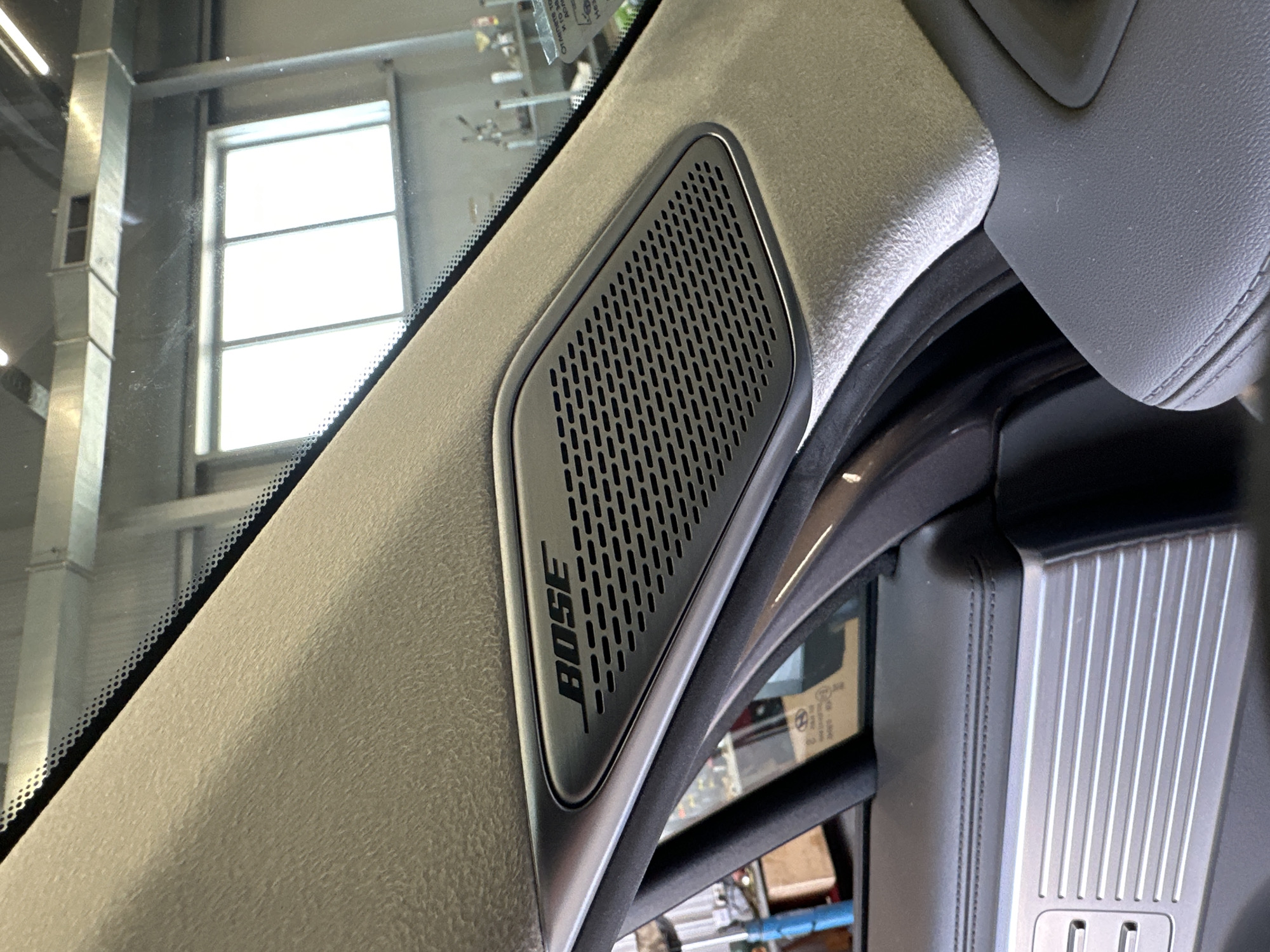 The image shows a close-up view of a Bose speaker grille integrated into the A-pillar of a car interior. The speaker grille is a dark, elongated hexagonal shape with a metallic border, and it features a pattern of small, rectangular perforations. The Bose logo is prominently displayed in black text above the perforations. The A-pillar itself is covered in a light-colored, textured fabric, likely suede or a similar material. Below the A-pillar and speaker, the image reveals a portion of the car's door panel, which appears to be made of dark leather with detailed stitching and a textured metallic accent piece. The background is out of focus, but suggests an indoor environment, possibly a garage or workshop, with a window, industrial shelving, and various objects. The lighting suggests ambient daylight coming from the window.