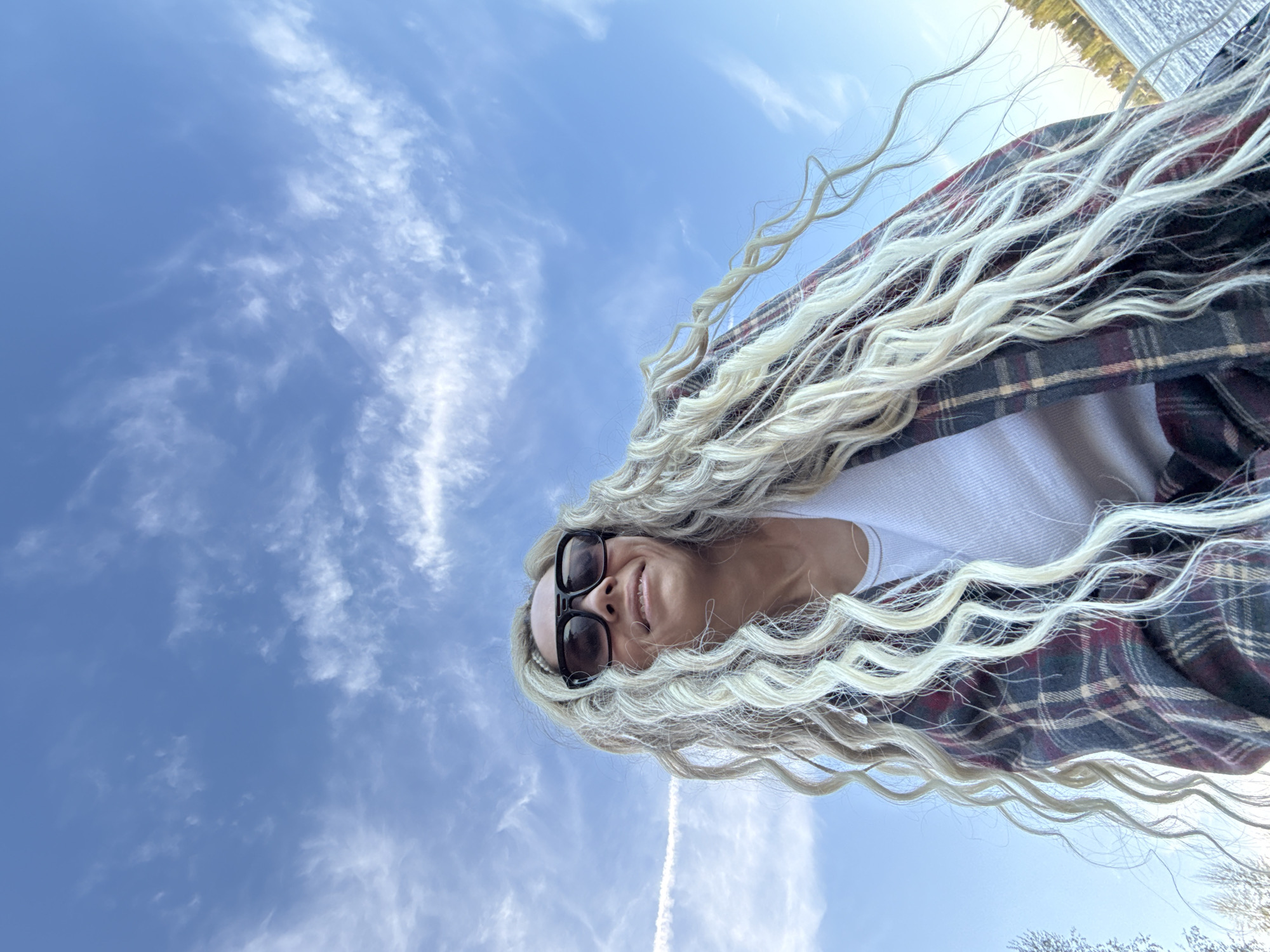A woman with blonde, wavy hair, wearing a white top and a dark plaid shirt, poses against a blue sky with scattered white clouds. She wears dark sunglasses and smiles slightly. The bottom edge of the frame includes bare tree branches, suggesting a natural, outdoor setting. The location is not identifiable as Rusanovo, Russia, based on the visual information in the image.