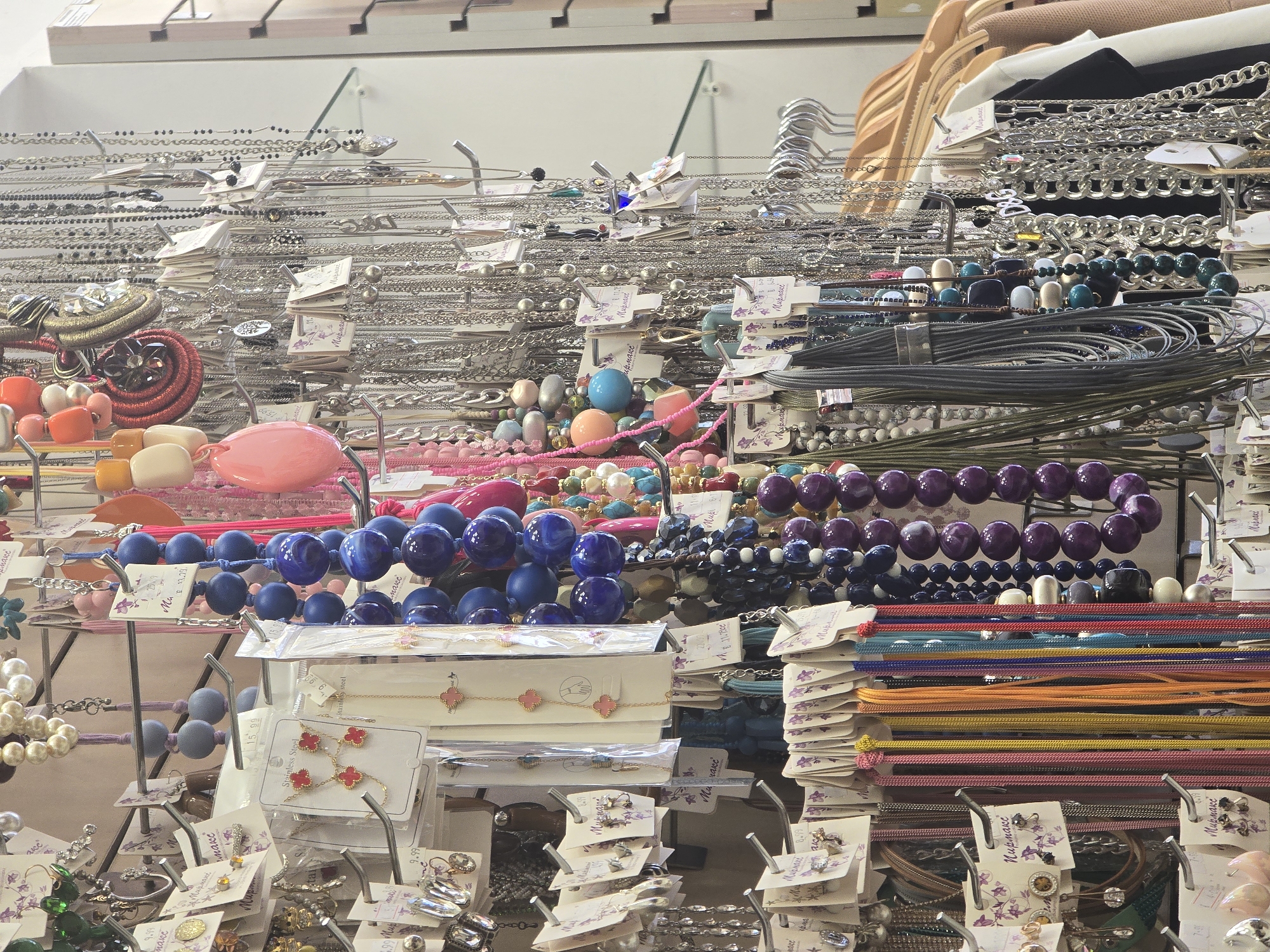 The image captures a detailed view of a densely packed retail display of fashion accessories within a Mirmaks store located in Sofia, Bulgaria. A wide assortment of necklaces and earrings is prominently featured, arranged on chrome-plated metal display racks equipped with numerous hooks. Necklaces vary in style, including long beaded strands in shades of royal blue, deep purple, and various bright colors (pink, orange, green, multi-color), alongside numerous silver-toned and gold-toned metal chain necklaces, some with small pendants. Earrings are displayed on individual white cards, often labeled "Stainless Steel," with many cards bearing the Cyrillic brand name "Мирмакс" and numerical prices such as 9.99, 13.99, 15.99, and 22.00. The background shows a light-colored wall, hints of glass shelving, and to the far right, a partial clothing rack with wooden hangers supporting plain garments in black, white, and beige. No individuals are visible in the scene.