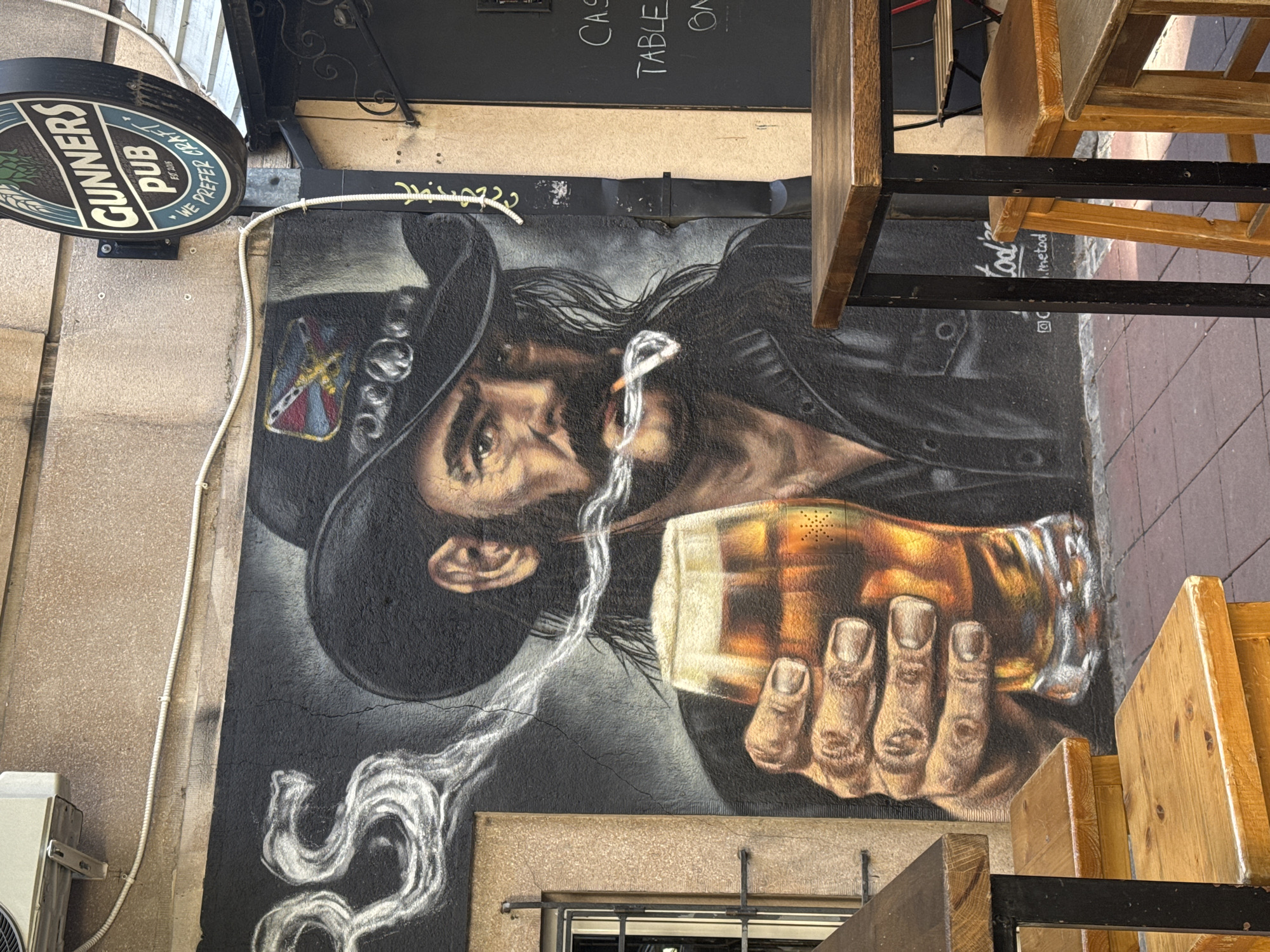 This media file captures a vibrant mural painted on an exterior wall, likely in Belgrade, Serbia. The mural depicts a close-up of a man's face and upper body, rendered in a realistic yet stylized manner. He wears a black cowboy hat and a dark jacket. A cigarette dangles from his lips, with smoke swirling around his face. In his right hand, he holds a glass of amber liquid, likely beer or whiskey, filled with ice. The man's expression is intense and perhaps a little weary, with a keen gaze.
To the left of the mural, a circular sign for "GUNNERS PUB EST. 2015 WE PREFER CRAFT" is visible, suggesting the mural is a decoration for the establishment. Above the mural, blackboards with handwritten text are partially visible, with words like "CAS", "TABLE", and "ON" discernible, possibly indicating menu items or seating arrangements. Wooden chairs and tables are positioned in the foreground and to the right of the image, suggesting an outdoor seating area for the pub. The scene is illuminated by natural light, and the overall ambiance feels casual and urban, hinting at a lively pub environment. The cracks in the wall beneath the mural indicate its age and exposure to the elements.