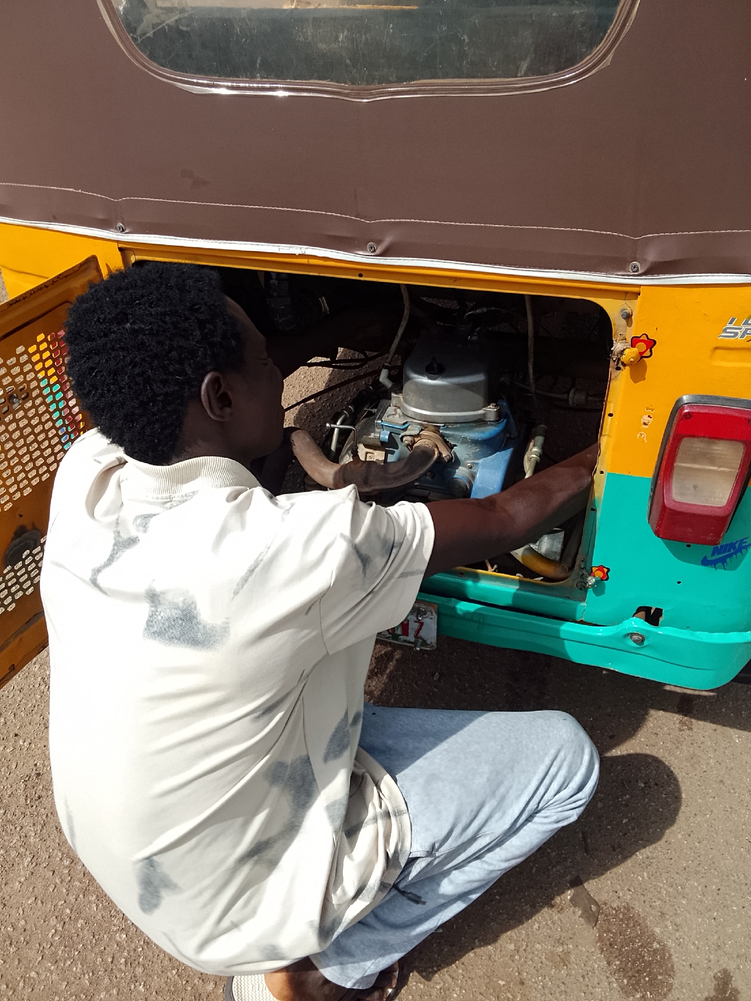 A man is kneeling and working on the engine of a yellow and teal auto rickshaw. The rickshaw's engine compartment is open, and the man's arms are reaching inside. He is wearing a light-colored t-shirt with darker markings and light-colored pants. The rickshaw has a brown roof and an open door. The vehicle's taillight, with the Nike logo, is visible on the right side. The scene is taking place outside on a paved surface, presumably in Bukuru, Nigeria, as requested. The man's shadow is cast on the ground.