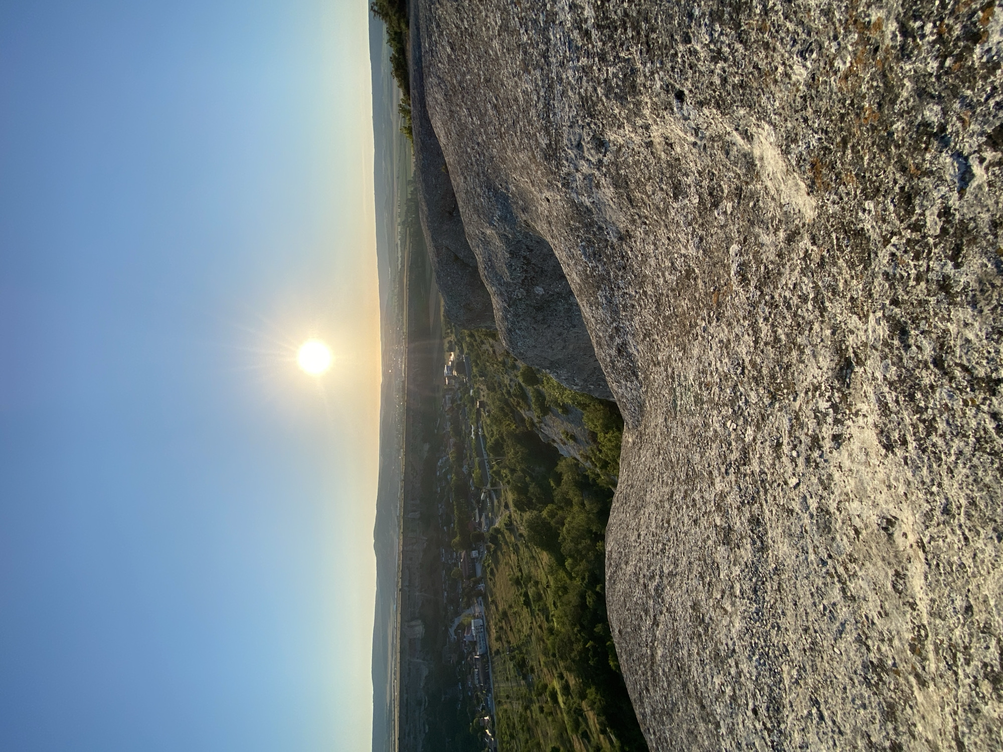 The image depicts a panoramic view of a valley with a settlement in the distance, bathed in the warm light of a setting sun. The foreground is dominated by a rough, rocky surface with a granular texture, likely sandstone, which appears weathered and possibly eroded. This rocky outcrop creates a natural viewpoint looking down into the valley.
In the middle ground, a small town or village is visible, characterized by clusters of buildings with red and white roofs, interspersed with patches of lush green vegetation. The town is situated within a valley or depression, bordered by steep, rocky cliffs or escarpments on one side. The surrounding landscape is a mix of cultivated fields and dense green foliage, suggesting a rural or semi-rural environment.
The background features rolling hills and distant mountains under a clear, bright blue sky. The sun, a prominent circular orb emitting a strong glare, is positioned in the upper center of the frame, indicating it is either sunrise or sunset. The sky transitions from a pale yellow near the horizon to a deep blue overhead.
The specific location provided, Красномакское сельское поселение, Ukraine, suggests this is a landscape view from a high vantage point overlooking a settlement within this region. The scene is devoid of any people or animals, focusing solely on the natural landscape and the human settlement integrated within it. The overall impression is one of natural beauty and tranquility, captured during the golden hour.