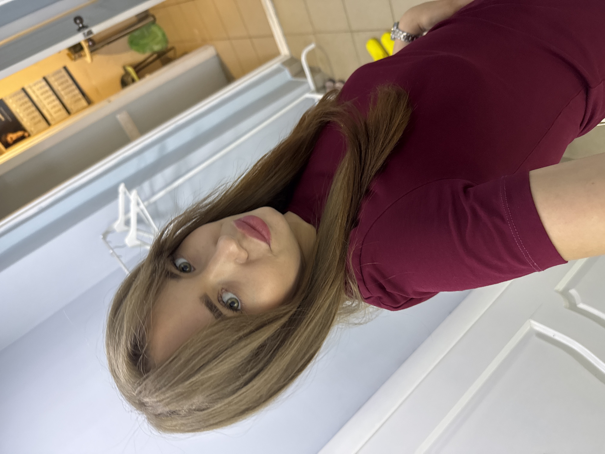 The image depicts a woman in her late 20s to early 30s, with long, straight, light brown hair and green eyes. She is wearing a dark red, short-sleeved dress and a silver-toned wristwatch on her left wrist. Her makeup includes visible lipstick. The woman is taking a selfie, as indicated by her pose and the angle of the shot.
The background suggests an indoor setting, possibly a salon or a changing room. To the left, there are white doors with decorative paneling. In the center and to the right, a hallway is visible. It is brightly lit with overhead lighting, and the walls are painted a light blue. Further down the hallway, a door is ajar, revealing a room with tiled flooring and yellow chairs. A green trash bag is also visible in this room. A rack with several white hangers is present in the background, to the left of the hallway.
The image does not contain any explicit information that would allow for pinpointing the exact location within the city of Chaikovsky, Russia, beyond the assumption that it is an indoor environment.
