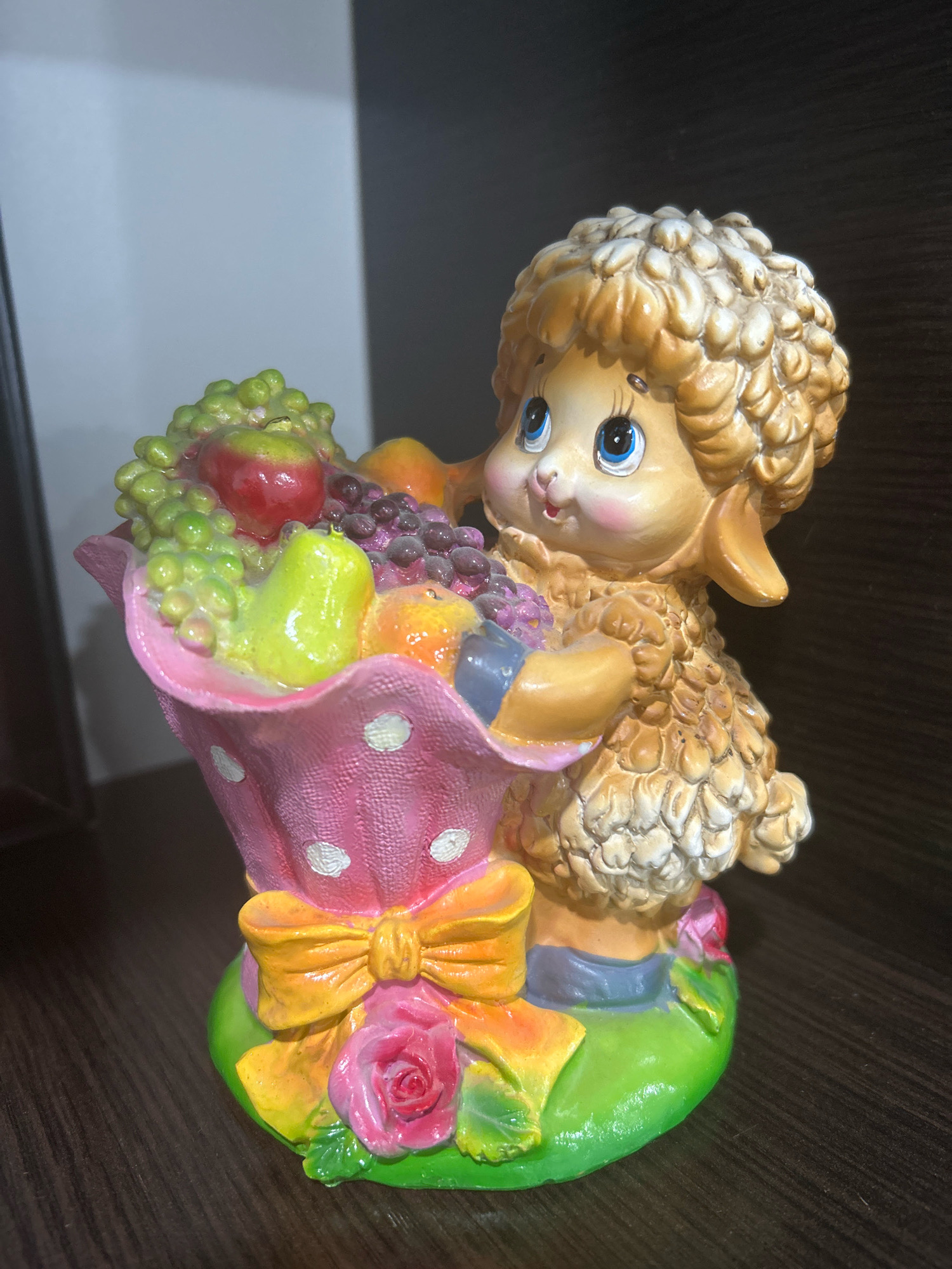 This is a close-up, eye-level, indoor shot of a ceramic figurine of a sheep carrying a basket of fruit. The figurine is detailed, with a fluffy wool texture on the sheep and vibrant colors for the fruit and basket. The sheep has large blue eyes, rosy cheeks, and a gentle expression. The basket is pink with white polka dots and a yellow ribbon tied around it, adorned with a pink rose at the base. The fruit in the basket includes grapes, an apple, a pear, and what appear to be tangerines. The figurine stands on a green base that mimics grass. The background is dark and out of focus, suggesting the figurine is placed on a shelf or table. There is no discernible connection to the city of Novopokrovskaya, Russia, as the image lacks any geographical identifiers.